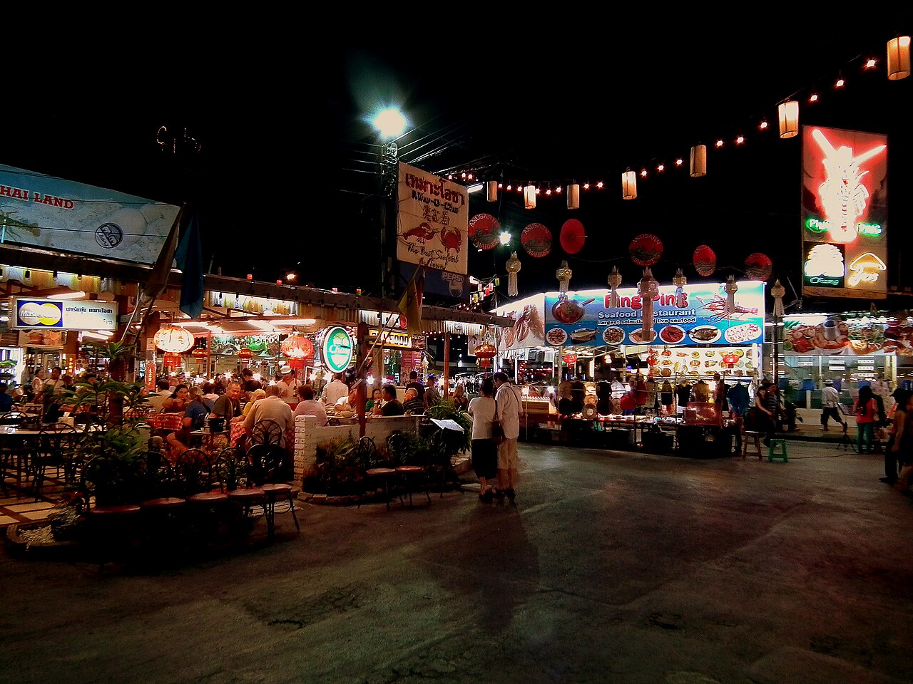 โซนอาหารกลางแจ้งของไนท์บาซาร์เชียงใหม่ยามค่ำ — ร้านอาหารทะเลย่างและร้านนั่งดื่ม โคมไฟแดงและไฟประดับห้อยเหนือลานนั่งกิน