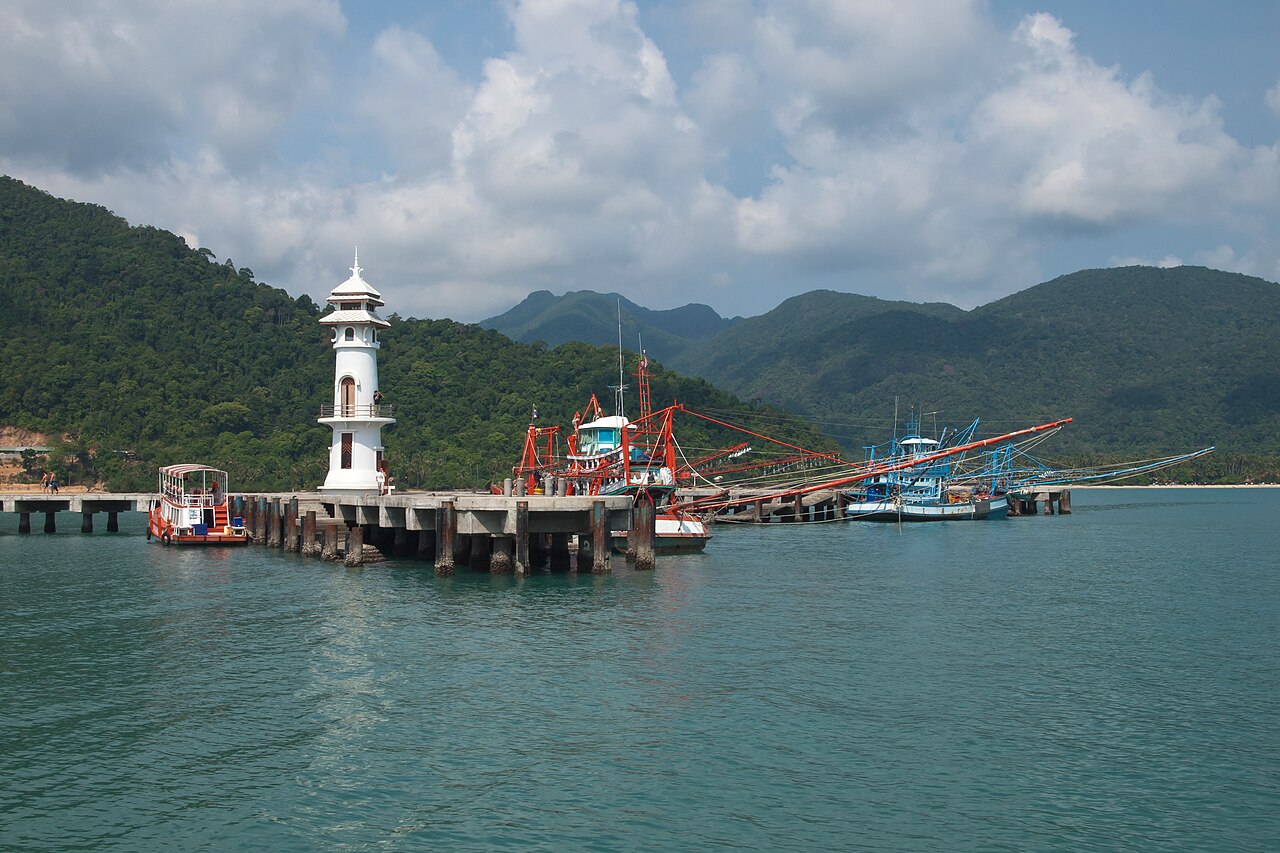 บางเบ้า เกาะช้าง — สะพานท่าเรือยื่นออกทะเล มีประภาคารสีขาวและเรือประมงสีสดจอดเรียง ภูเขาเขียวเป็นฉากหลัง