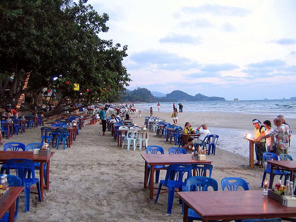หาดทรายขาว เกาะช้าง — แนวโต๊ะร้านอาหารริมหาดยามเย็น คนเดินเล่นบนทราย และเกาะเล็กๆ ในทะเลไกลออกไป