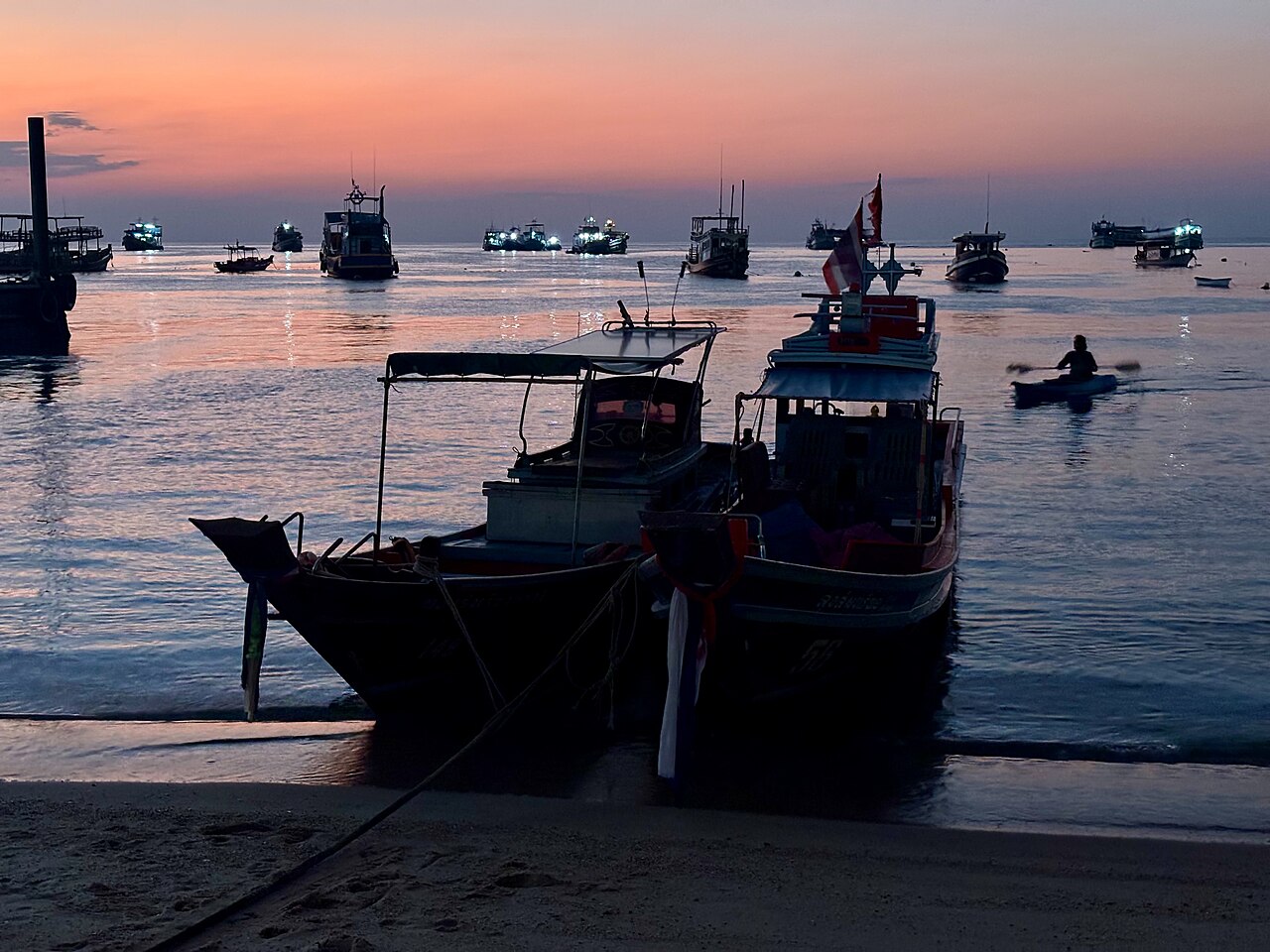 หาดทรายรีของเกาะเต่าช่วงพระอาทิตย์ตก กับเรือทอดสมออยู่หน้าหาดและท้องฟ้าสีส้มชมพู หาดหลักที่เป็นหัวใจของฉากคาเฟ่บนเกาะ