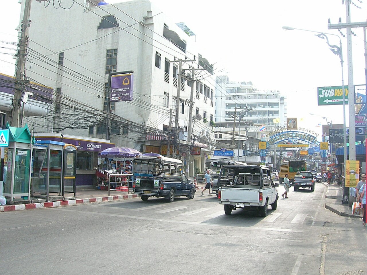 ถนนทางเข้าวอล์กกิ้งสตรีท พัทยา ตอนกลางวัน เห็นซุ้มป้ายทางเข้าไกล ๆ มีรถสองแถวสีฟ้าวิ่งบนถนนเลียบหาด