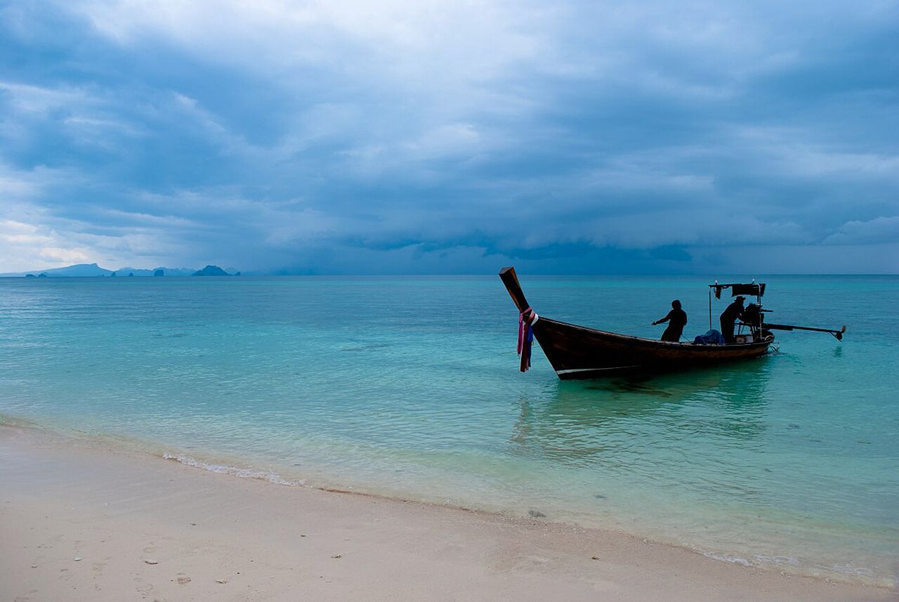 เกาะเฮ (เกาะคอรัล) ใกล้ภูเก็ต — หาดทรายขาวริมน้ำทะเลสีฟ้าเขียวใส มีเรือหางยาวจอดริมหาด ท้องฟ้าครึ้มเมฆ