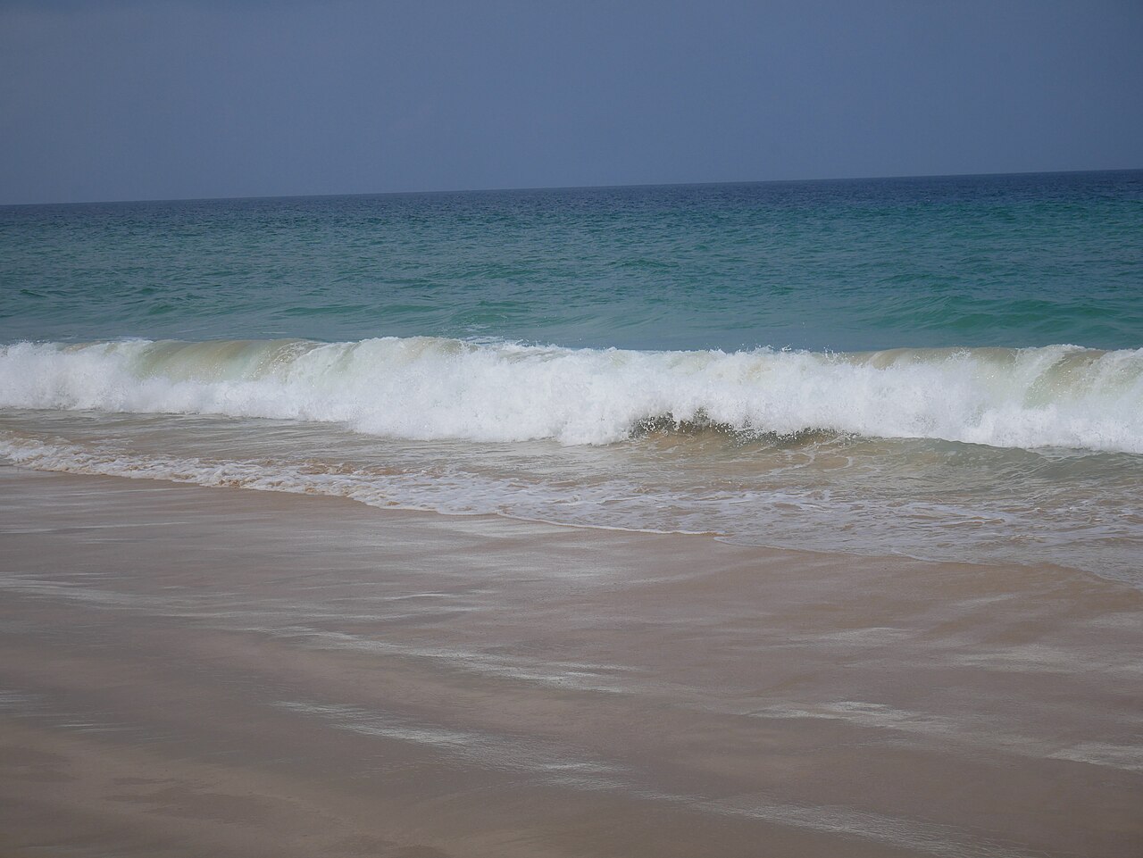 Karon Beach Phuket — a wave rolling onto a wide white-sand beach with turquoise sea meeting the sky