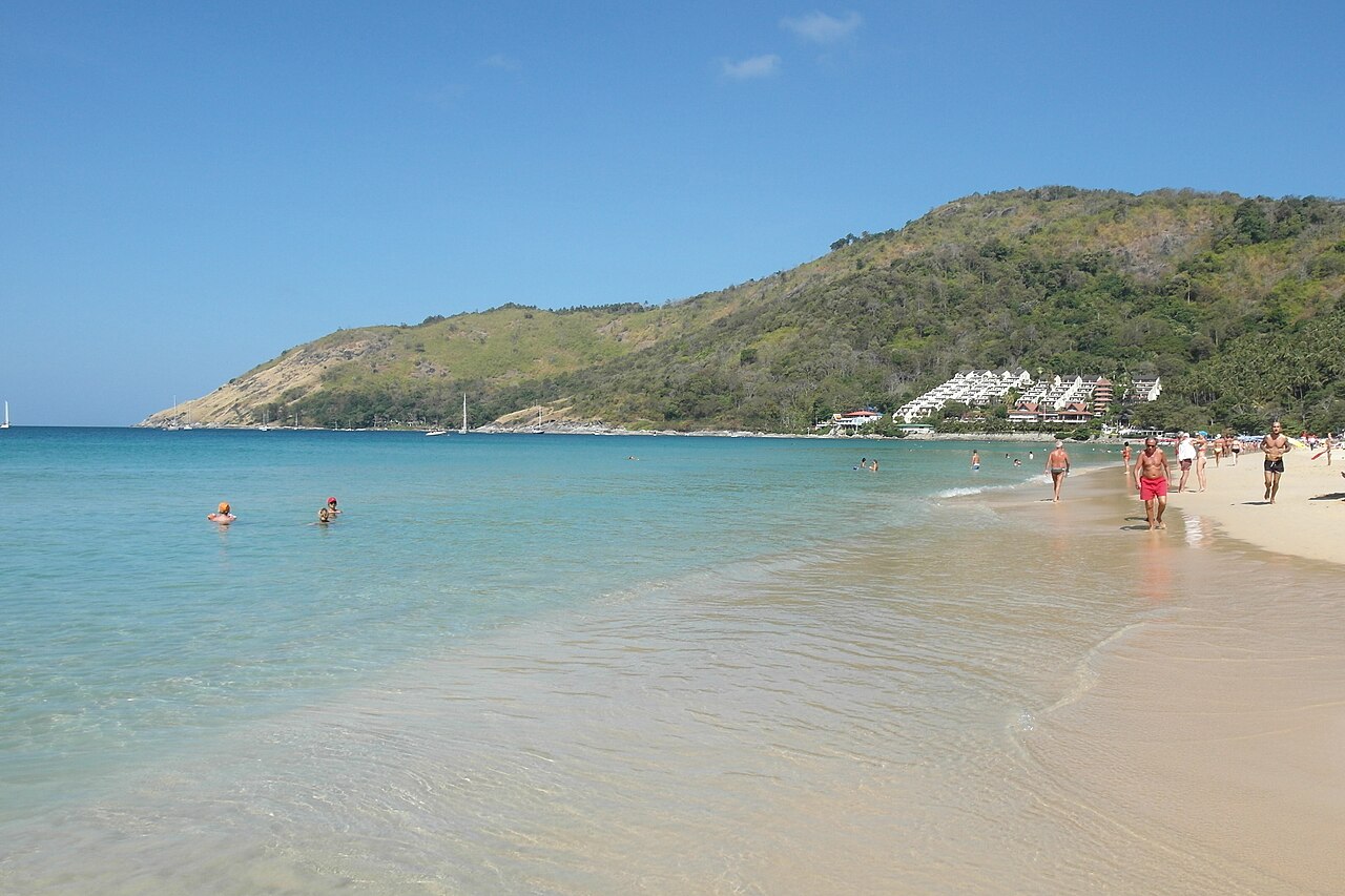 Nai Harn Beach, Phuket — a turquoise clear-water bay with white sand, green hills wrapping around it, a resort at the foot of the hill and people walking the shore