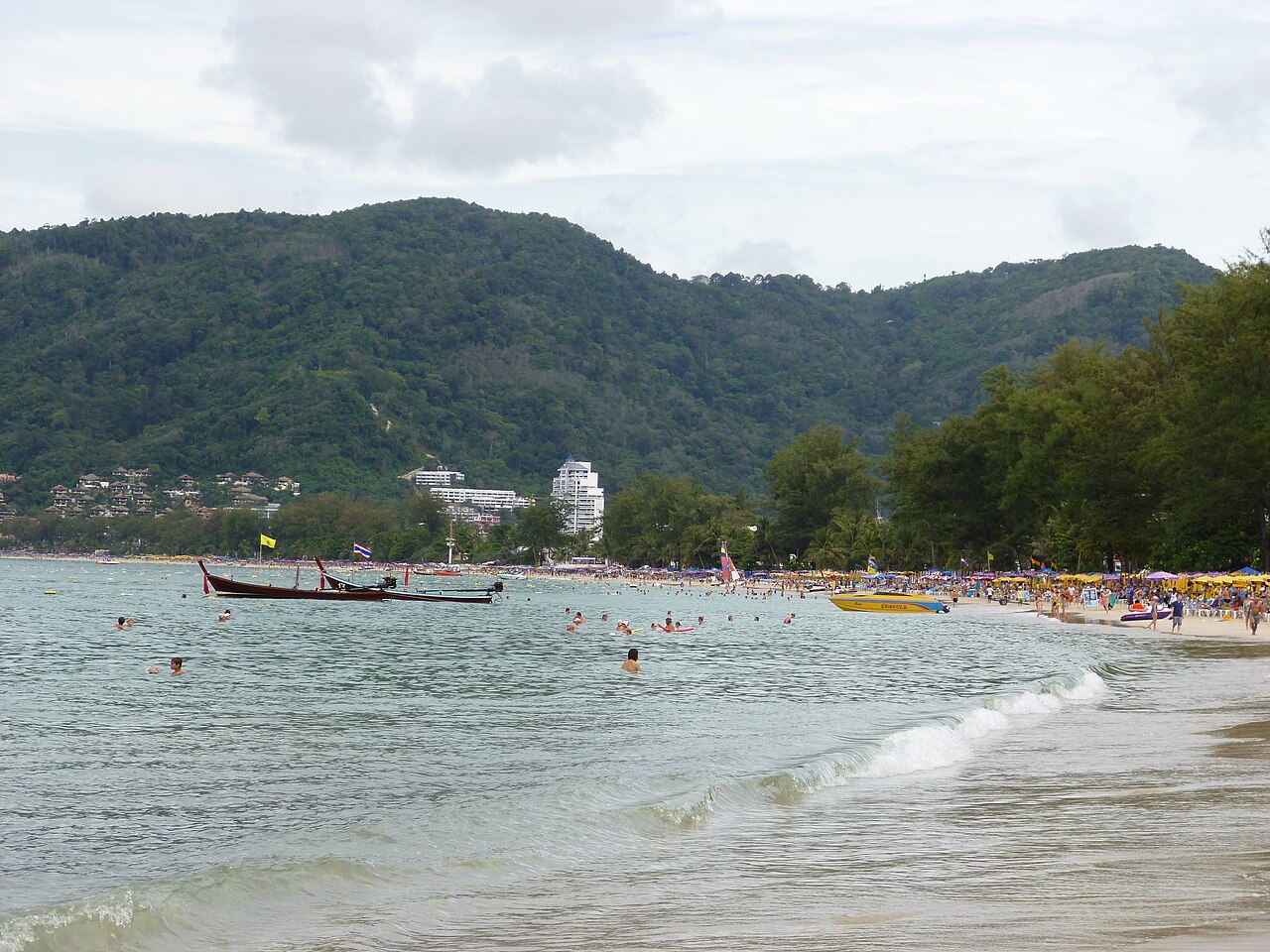 Patong Beach, Phuket — a busy curved beach with swimmers, long-tail boats moored offshore, and green hills and hotel towers behind