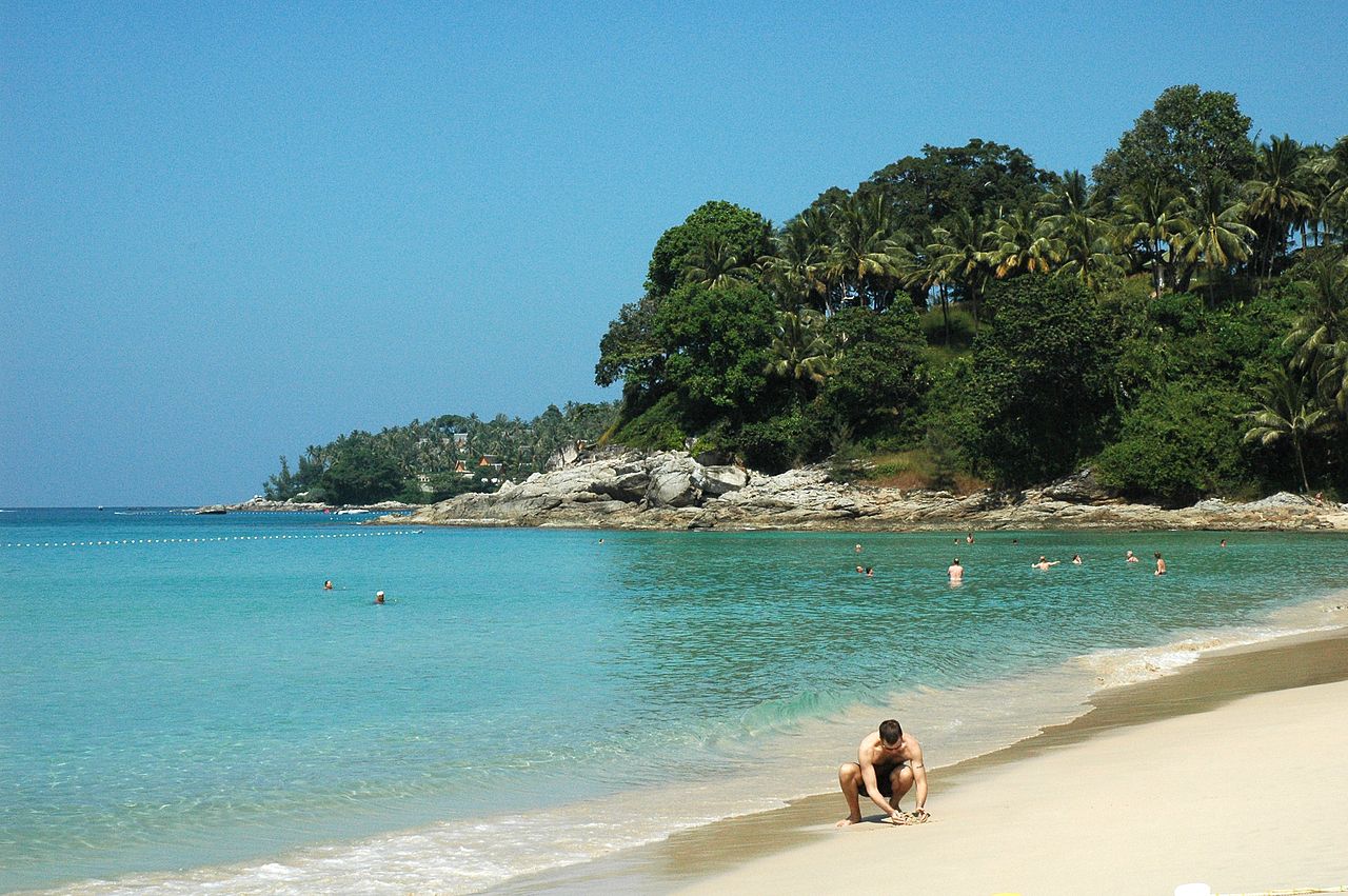 Surin Beach, Phuket — clear deep-blue water, white sand, a rocky headland and lush coconut palms, with swimmers behind a buoy line