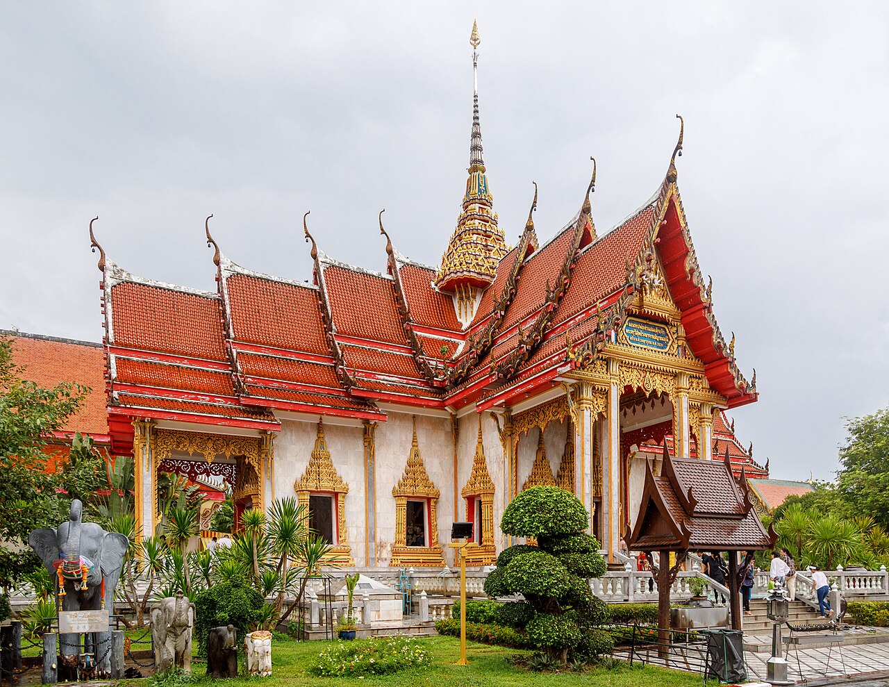 Wat Chalong Phuket — a Thai temple hall with a multi-tiered red-and-gold roof and a pointed finial, set among green gardens and elephant statues