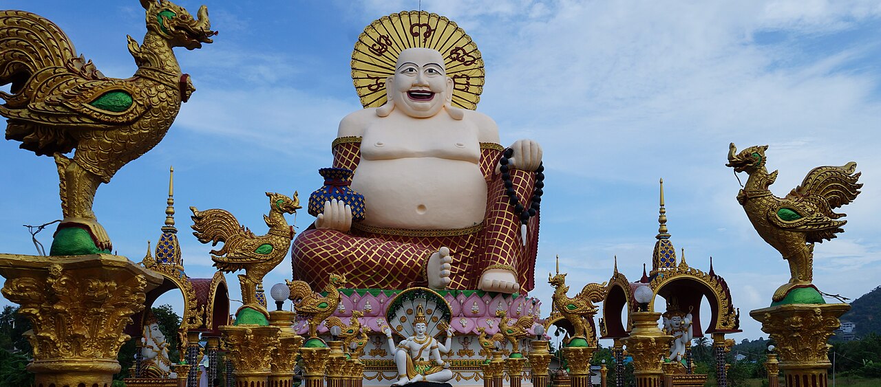 วัดปลายแหลม เกาะสมุย — พระสังกัจจายน์องค์ใหญ่และงานปูนปั้นสีทองวิจิตรตั้งอยู่กลางบึงน้ำ ท้องฟ้าสีฟ้าสดใส