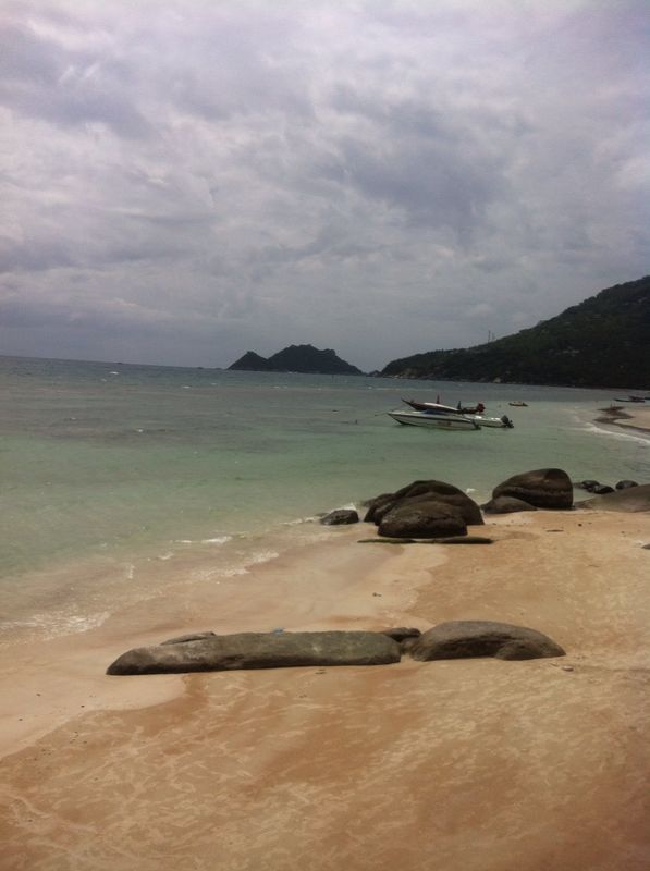 อ่าวเล็กในเกาะเต่า — ชายหาดทรายกับก้อนหินใหญ่ น้ำทะเลใส เรือเล็กจอด และเกาะเล็กลิบๆ ฉากหลัง