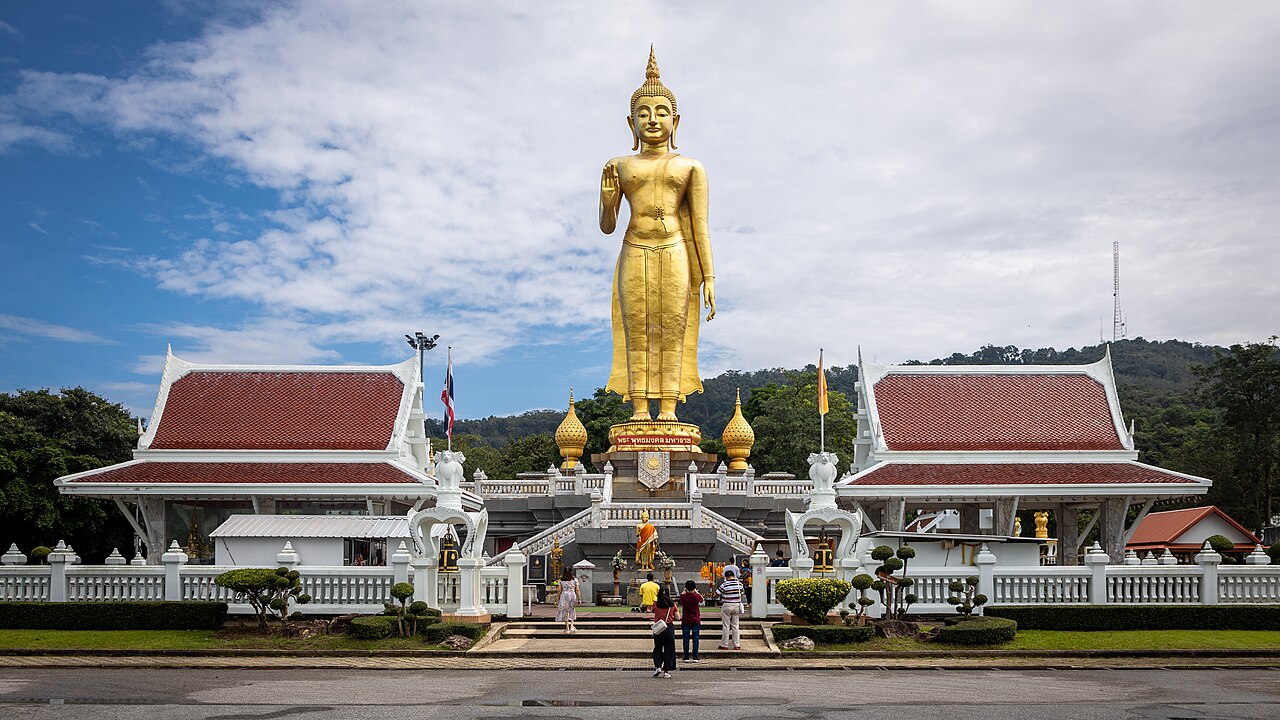 Phra Buddha Mongkol Maharaj, a giant golden standing Buddha on a hill at Hat Yai Municipal Park, flanked by white Thai pavilions with red roofs and a green hillside behind