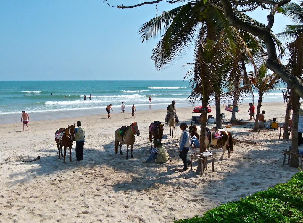 Hua Hin beach on the Gulf of Thailand by day, with ponies for hire along the shore and visitors walking on the sand under coconut palms