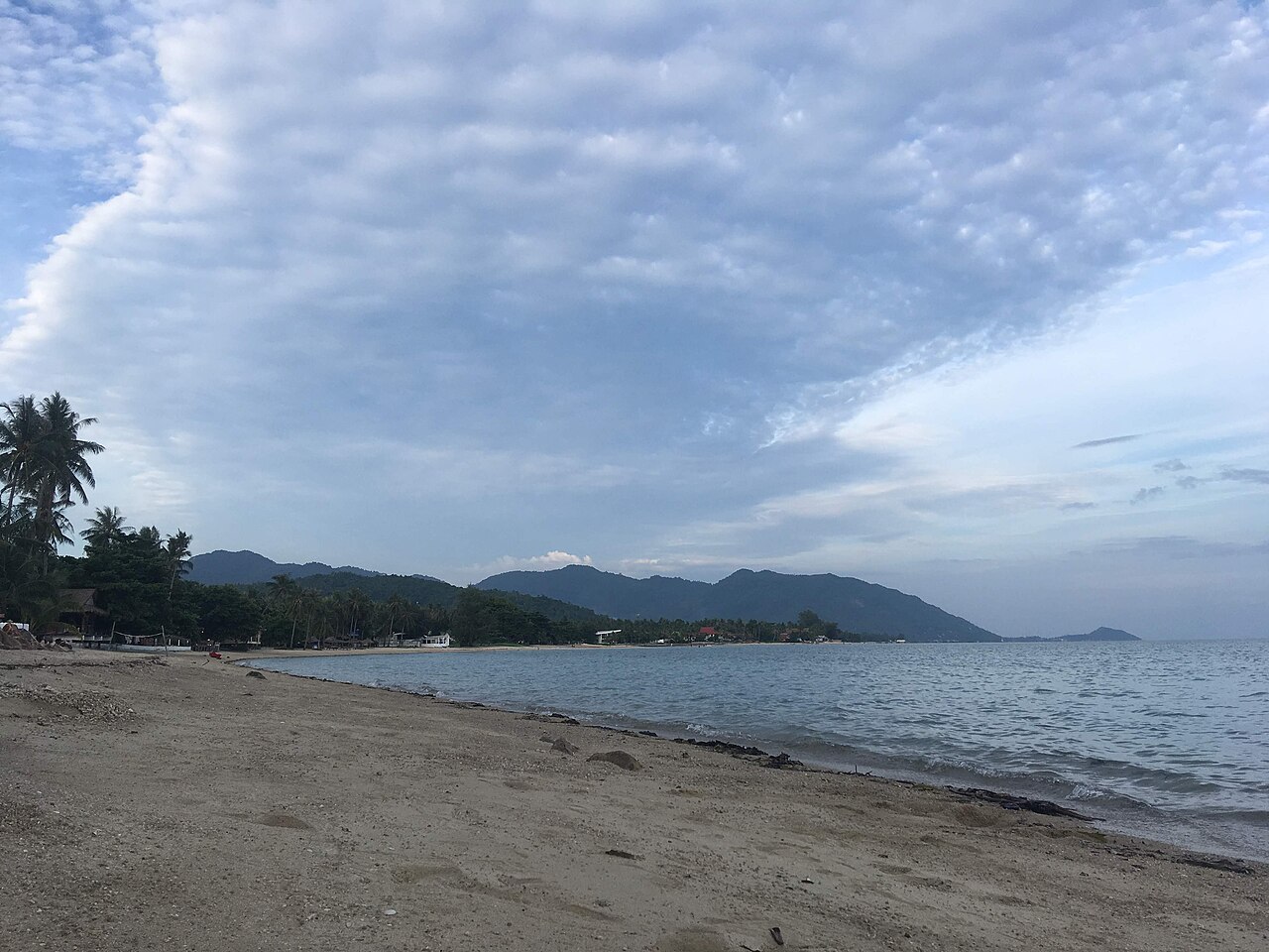 Baan Tai Beach, Koh Phangan — a long quiet beach at dusk with flat calm water, a line of mountains behind and a cloudy sky