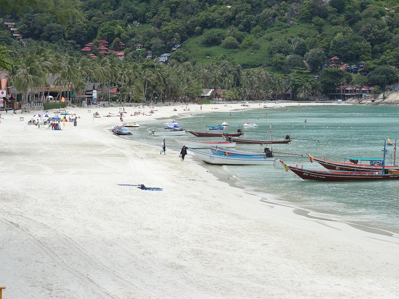 Haad Rin Beach, Koh Phangan — a long curve of white sand with longtail boats on emerald water and green hills behind