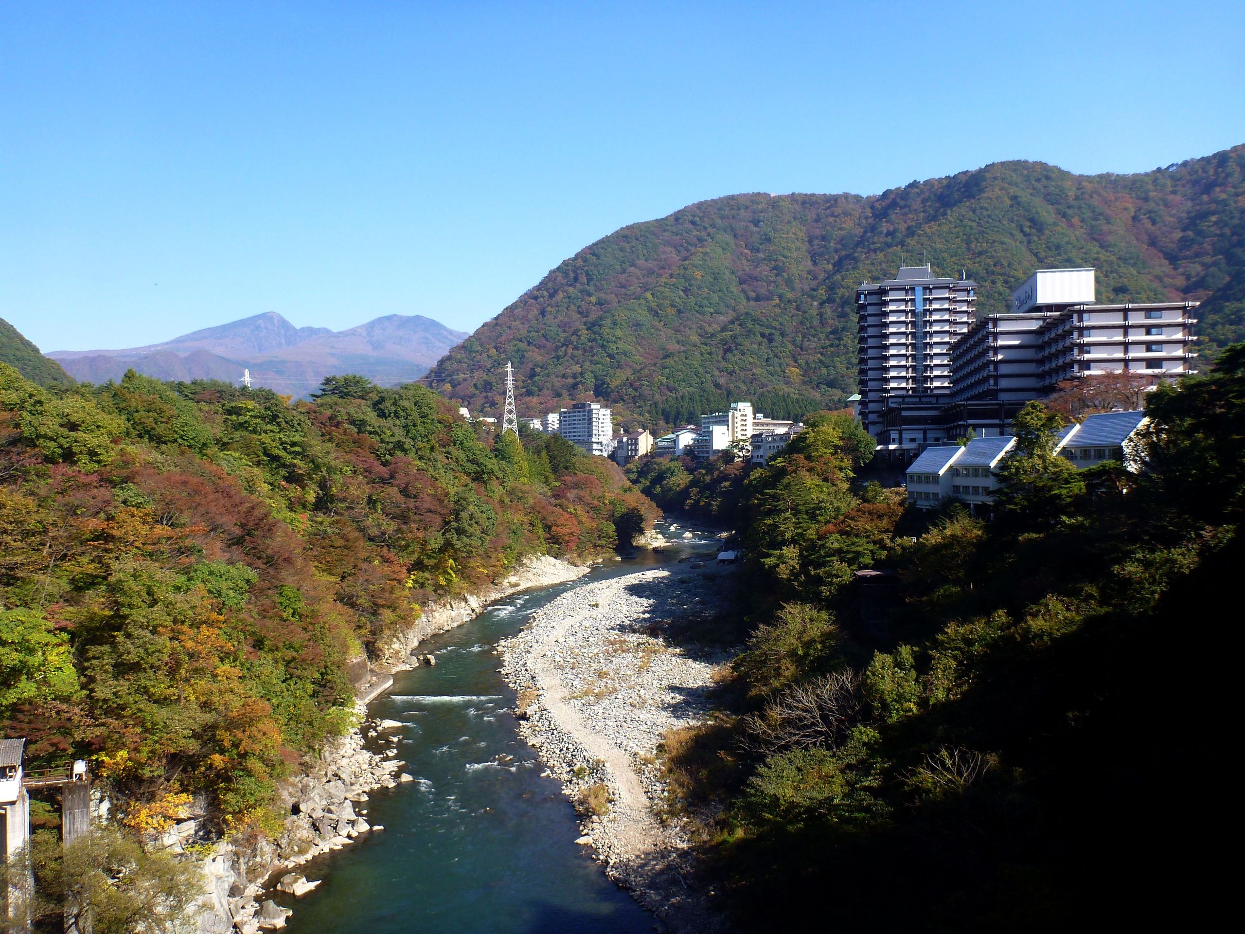 The Kinugawa River gorge at Kinugawa Onsen in autumn colours, large ryokan and resort hotels lining the cliff edge above a green river running through the valley