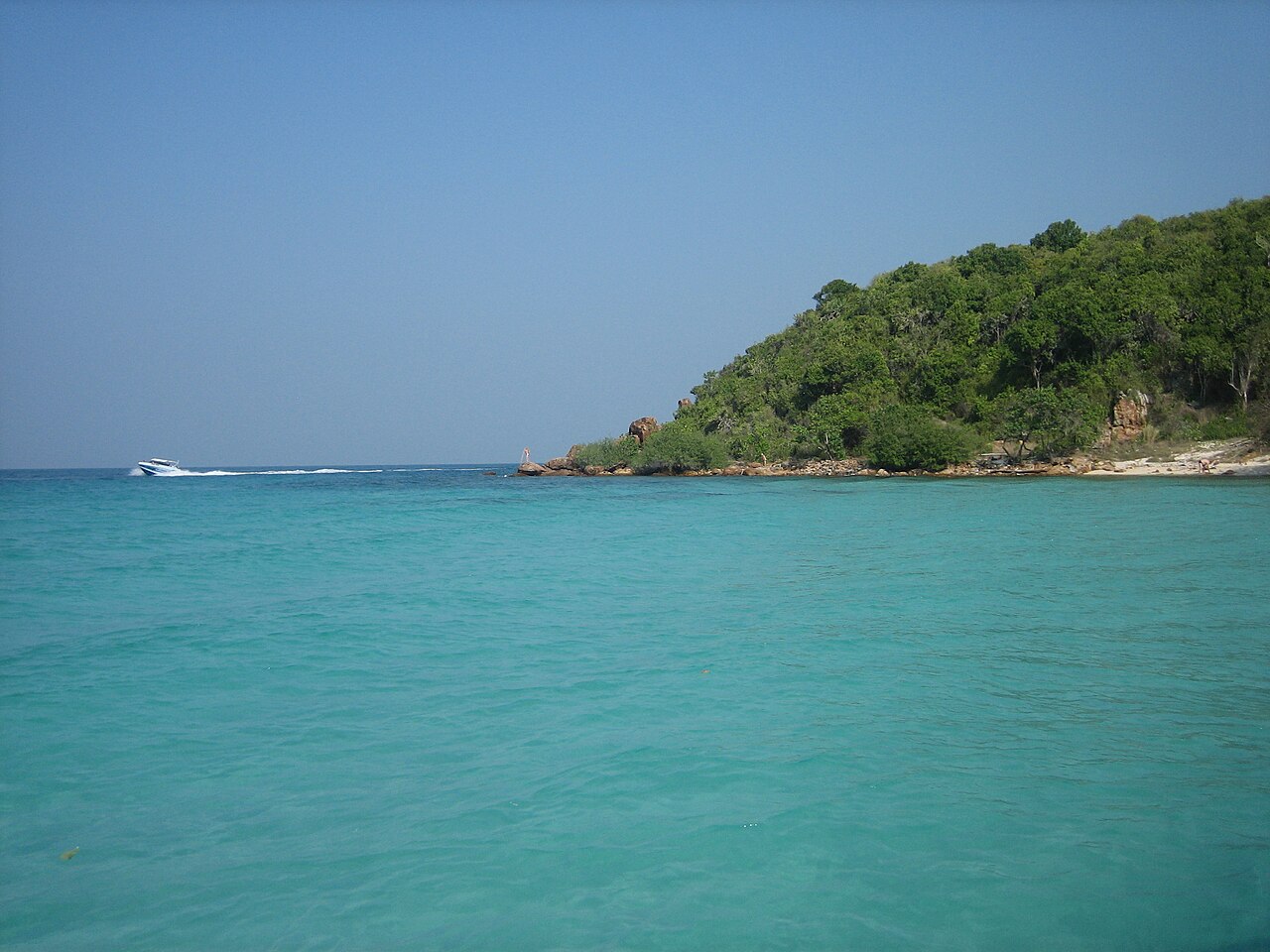 เกาะล้าน พัทยา — น้ำทะเลใสสีเขียวมรกตหน้าเกาะ มีแหลมหินและป่าเขียว เรือสปีดโบ๊ตแล่นผ่าน