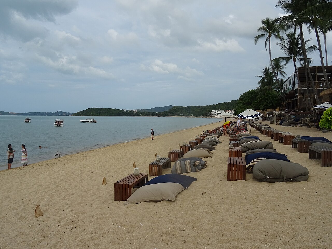Beanbag seats and low wooden tables of beachfront cafés and bars lined up on the sand of Bophut beach, Koh Samui, under coconut palms