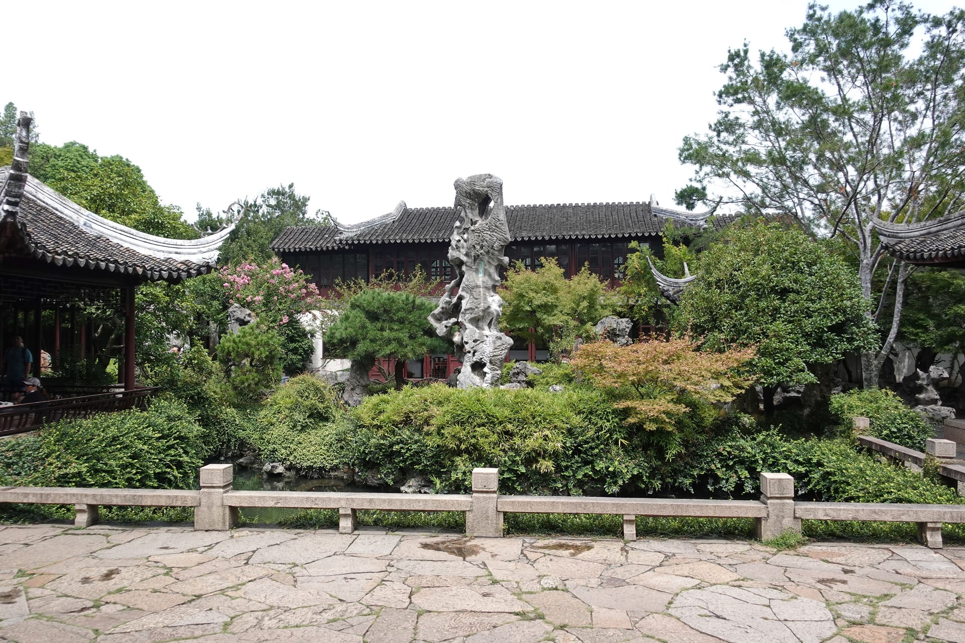 Lingering Garden, Suzhou — a flagstone courtyard with a tall vertical Taihu rock standing in the centre, dark-timber halls behind, greenery all around