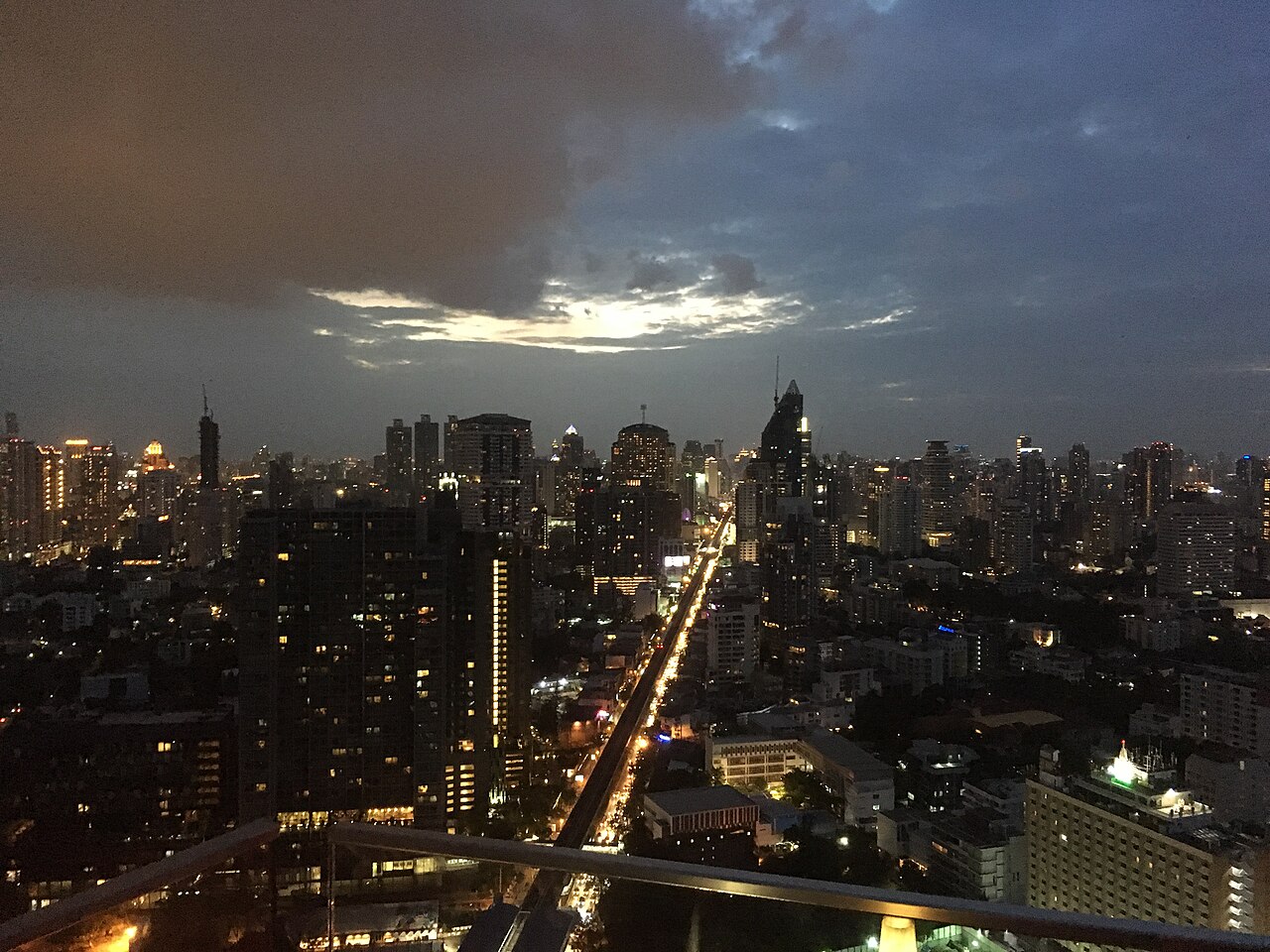 Sukhumvit Road at dusk seen from the glass railing of Octave on the 49th floor in Bangkok — the lit road cutting through a sea of towers under a post-rain sky