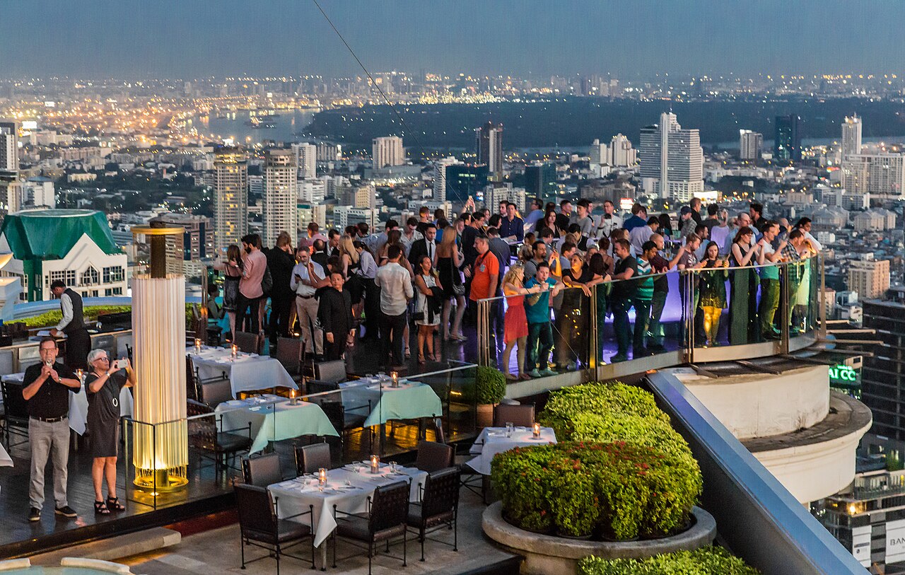 The Sky Bar terrace at lebua State Tower in Bangkok — guests at the 63rd-floor railing overlooking the Chao Phraya River at dusk, with Sirocco's dinner tables and a golden light column