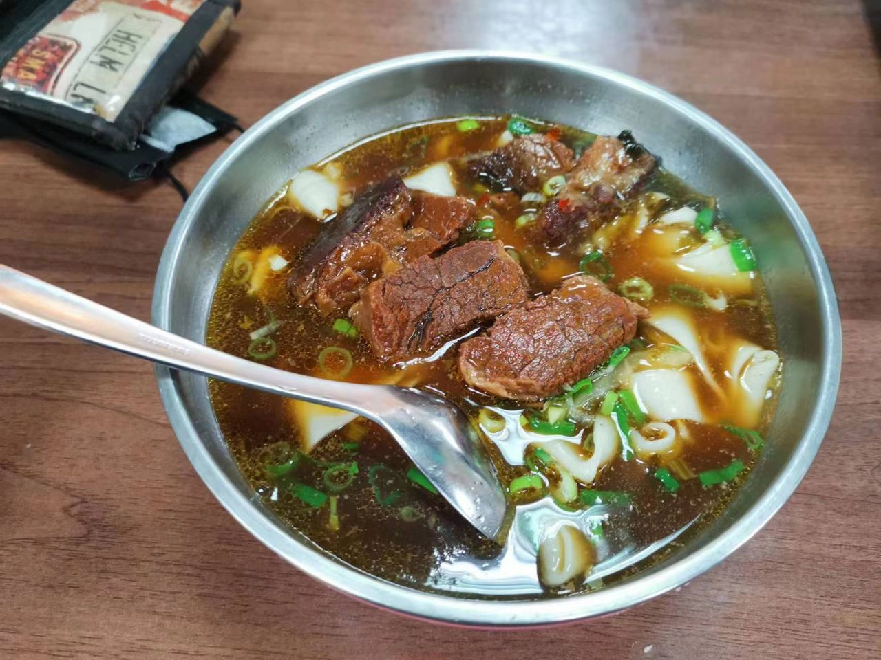 ก๋วยเตี๋ยวเนื้อน้ำซุปแดงหงเซาในชามสแตนเลส น้ำซุปสีน้ำตาลเข้มมีชิ้นเนื้อและต้นหอม