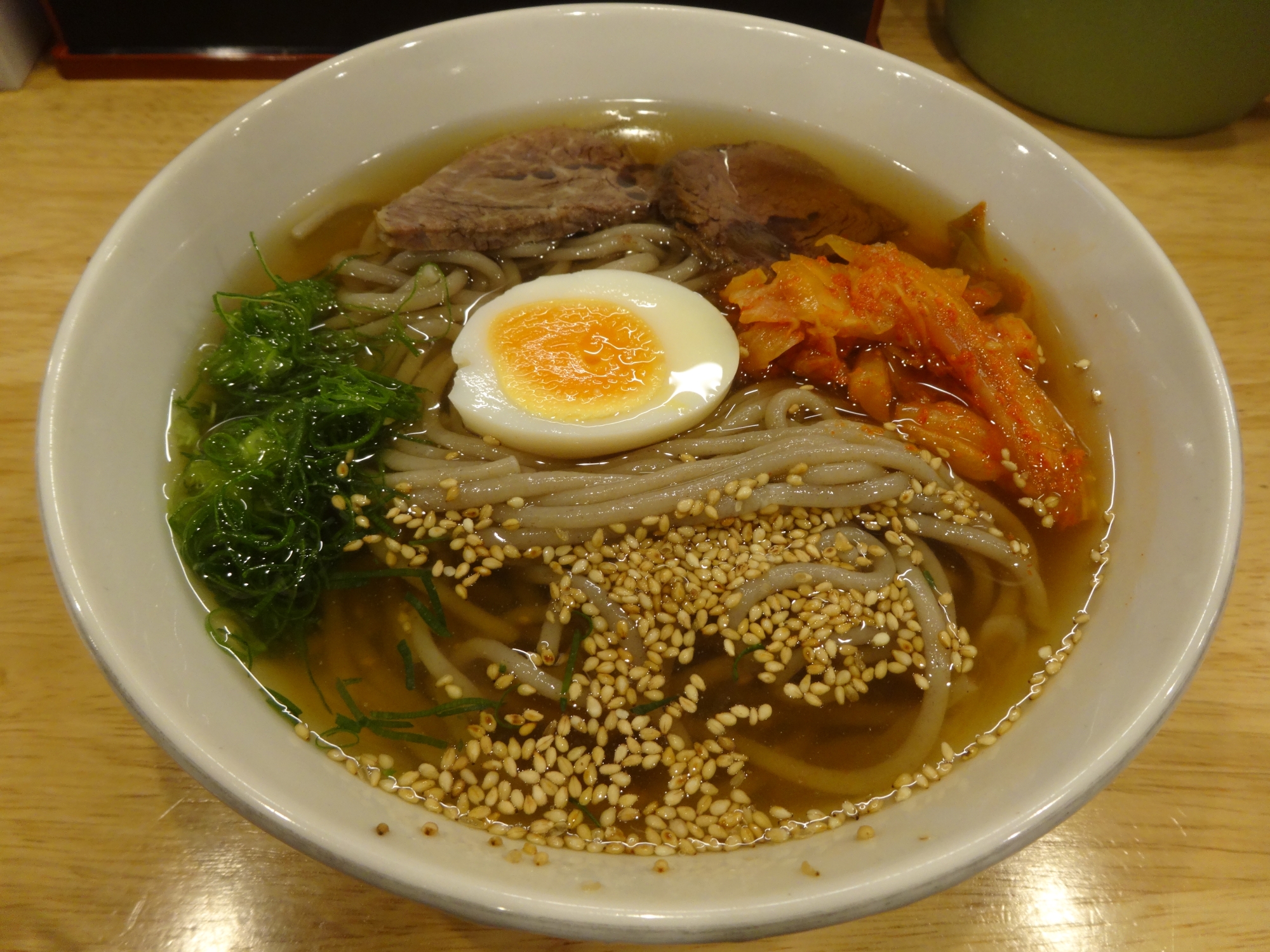 Beppu reimen cold noodles — a wide bowl of grey buckwheat noodles in clear chilled broth, topped with sliced beef, kimchi, a half-boiled egg and scattered sesame seeds