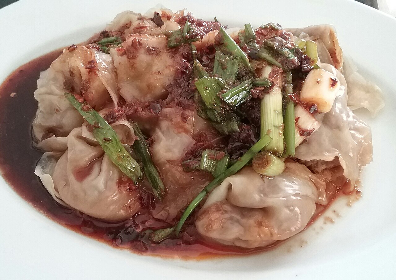 Long chao shou — folded Sichuan wontons on a white plate, glossy with red chili oil and scattered with scallion and ground Sichuan peppercorn
