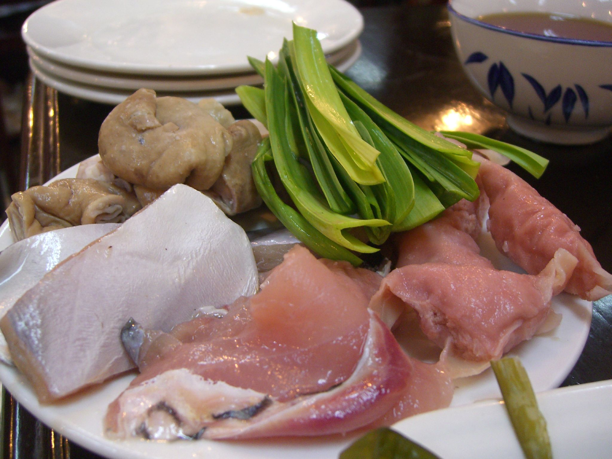 A plate of raw hotpot ingredients — thin sliced meats, intestine, fish fillets and garlic shoots, ready to cook