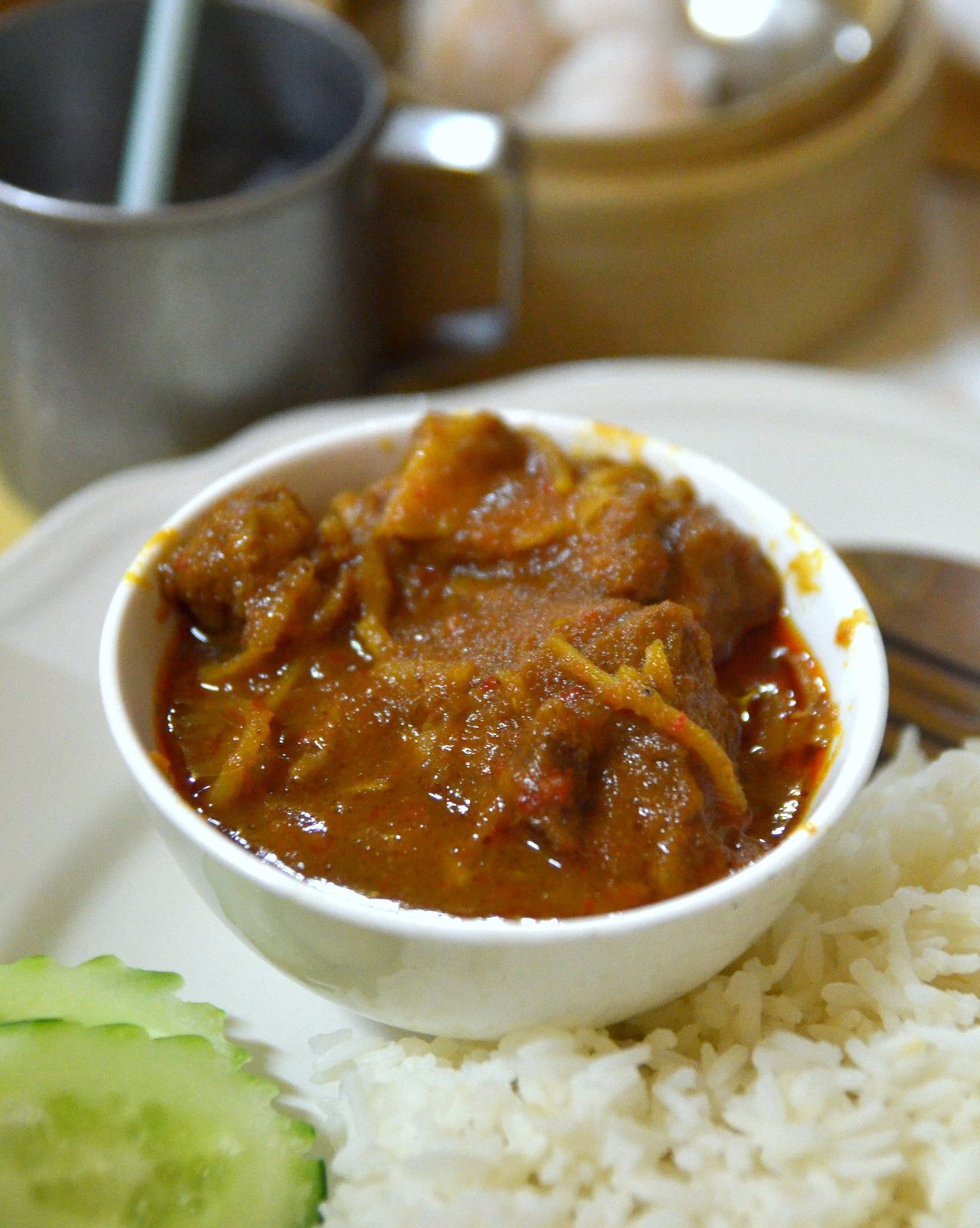 Gaeng hung lay — a thick reddish-brown pork belly curry with shredded ginger in a white bowl, served with rice and cucumber
