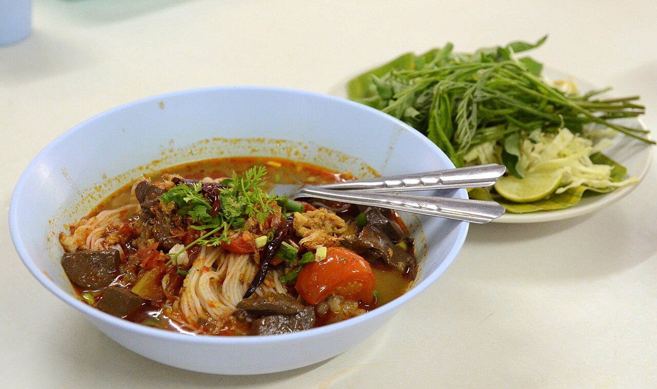 ขนมจีนน้ำเงี้ยวในชาม น้ำซุปสีแดงส้มมีมะเขือเทศและเลือดหมู เสิร์ฟคู่จานผักสดและกะหล่ำปลี