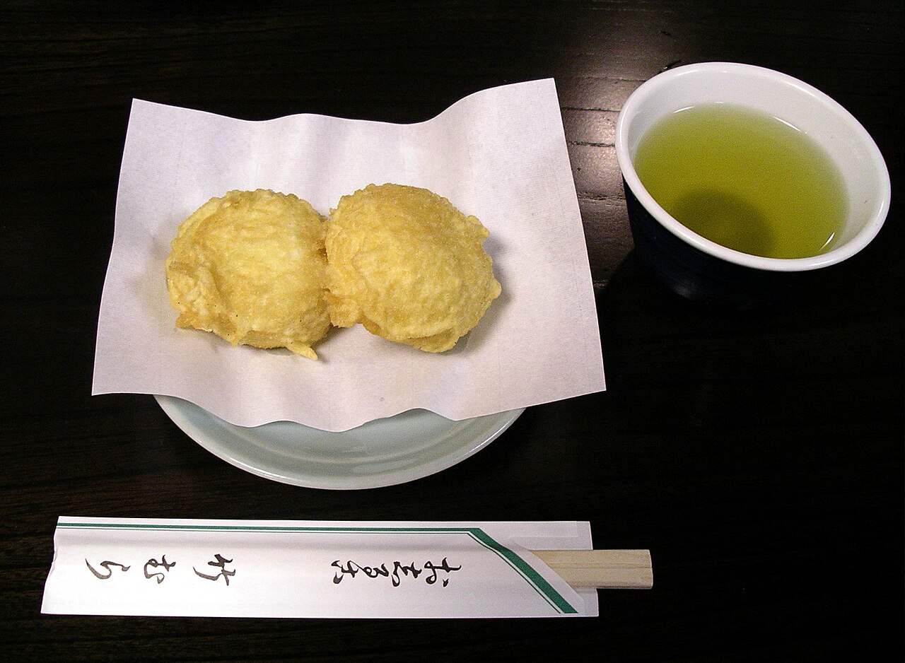 Two golden steamed-and-fried manju buns on white paper beside a cup of green tea — the traditional sweet snack sold in hot-spring towns like Hakone