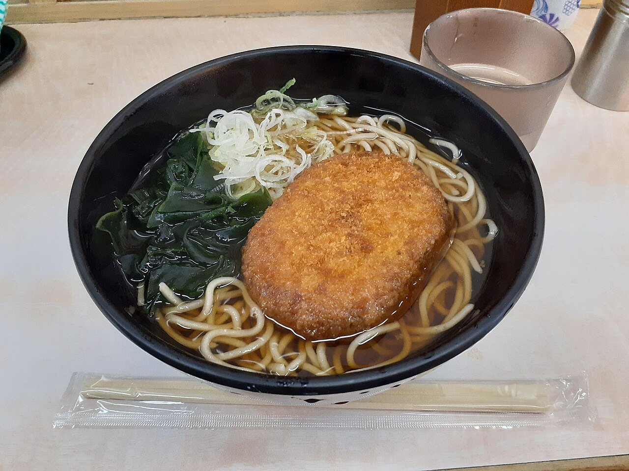 A bowl of Hakone soba in dark broth with wakame seaweed, sliced spring onion and a crisp fried korokke croquette on top, with chopsticks and a cup of tea