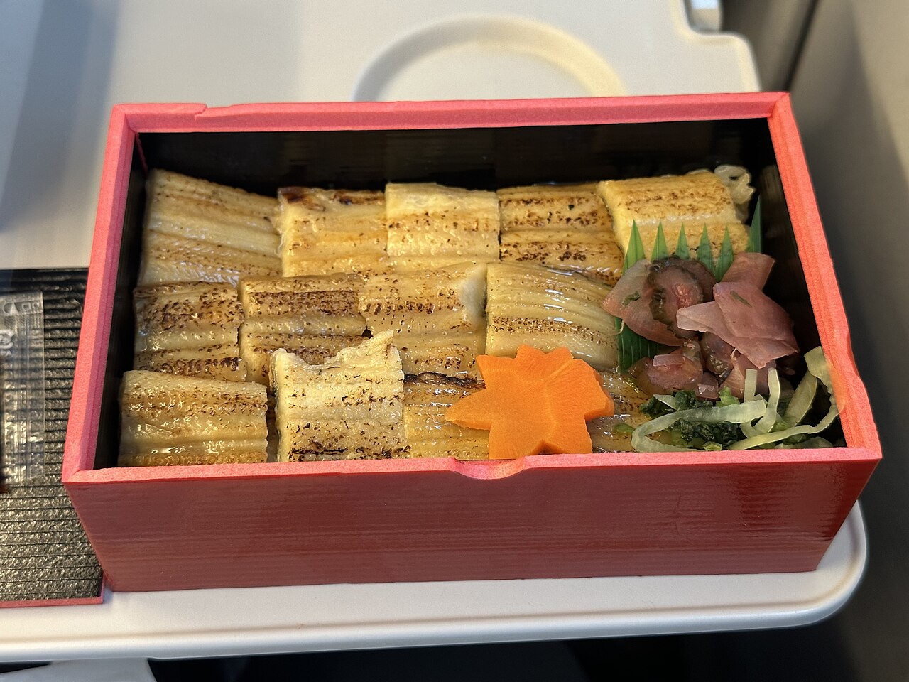 Anago-meshi bento box — a red lacquer box filled with sliced grilled conger eel glazed in sweet soy sauce, arranged over steamed rice, garnished with a carved carrot and green shiso