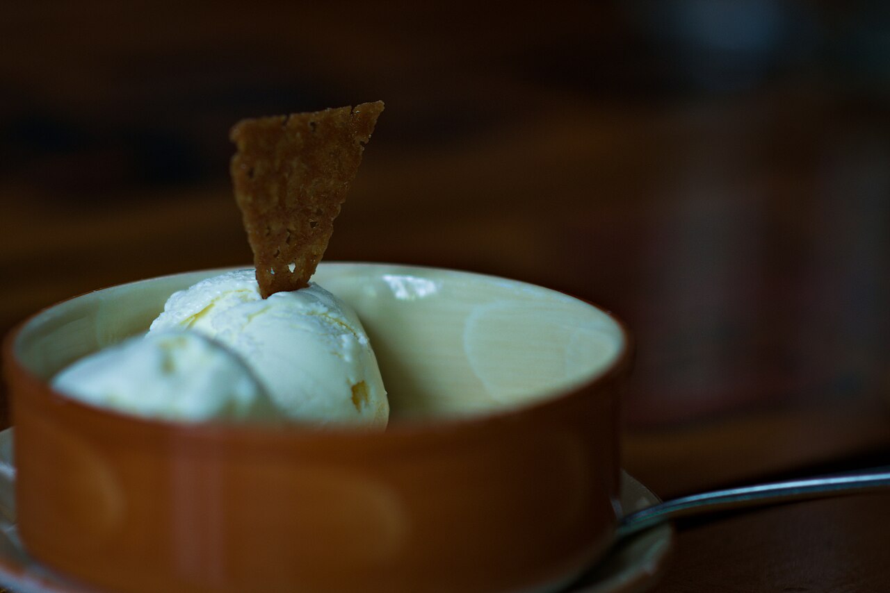 A scoop of cream-coloured fresh-milk ice cream in an orange wooden bowl, topped with a crisp cookie wafer, on a saucer with a spoon, Khao Yai farm café