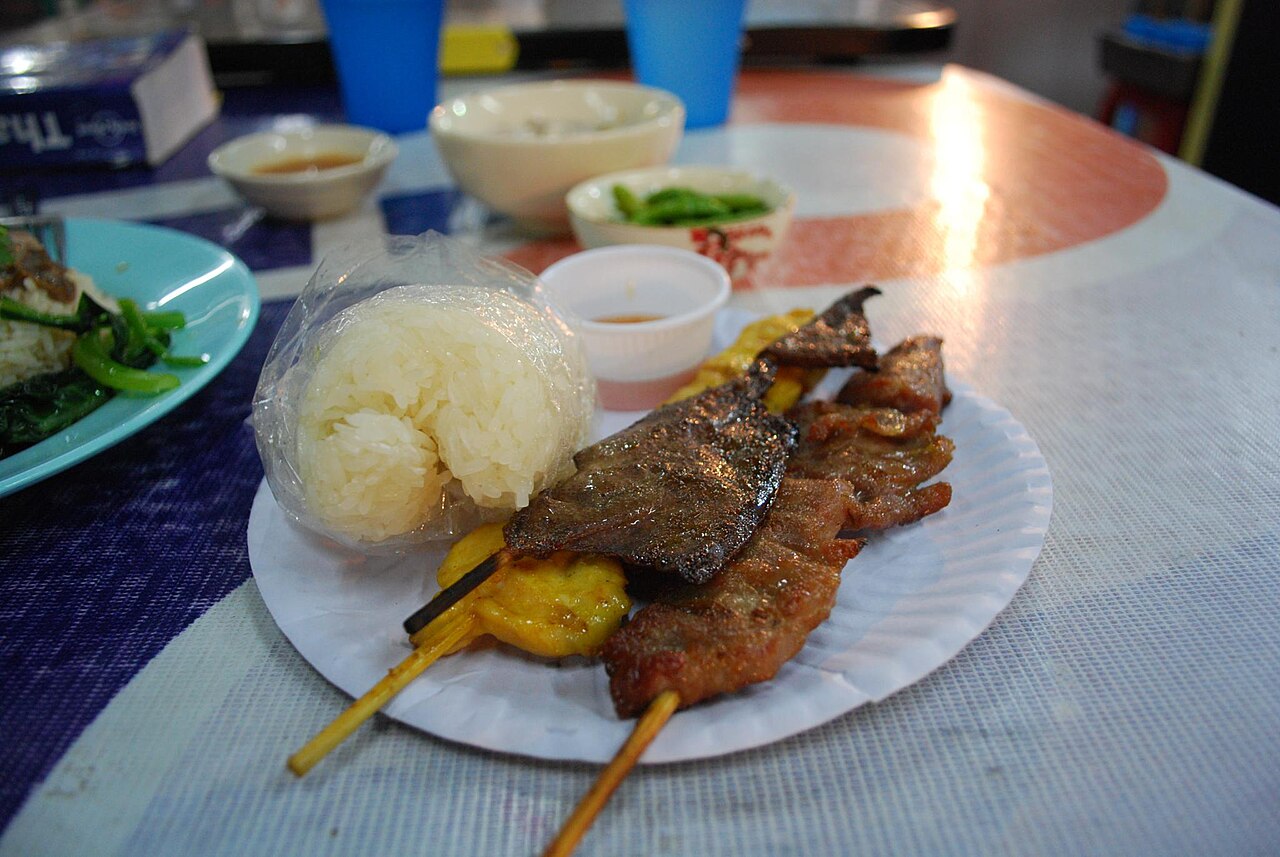 หมูปิ้งและเนื้อย่างเสียบไม้วางบนจานกระดาษคู่ข้าวเหนียวในถุง อาหารริมทางแบบที่เจอในตลาดเย็นปากช่อง