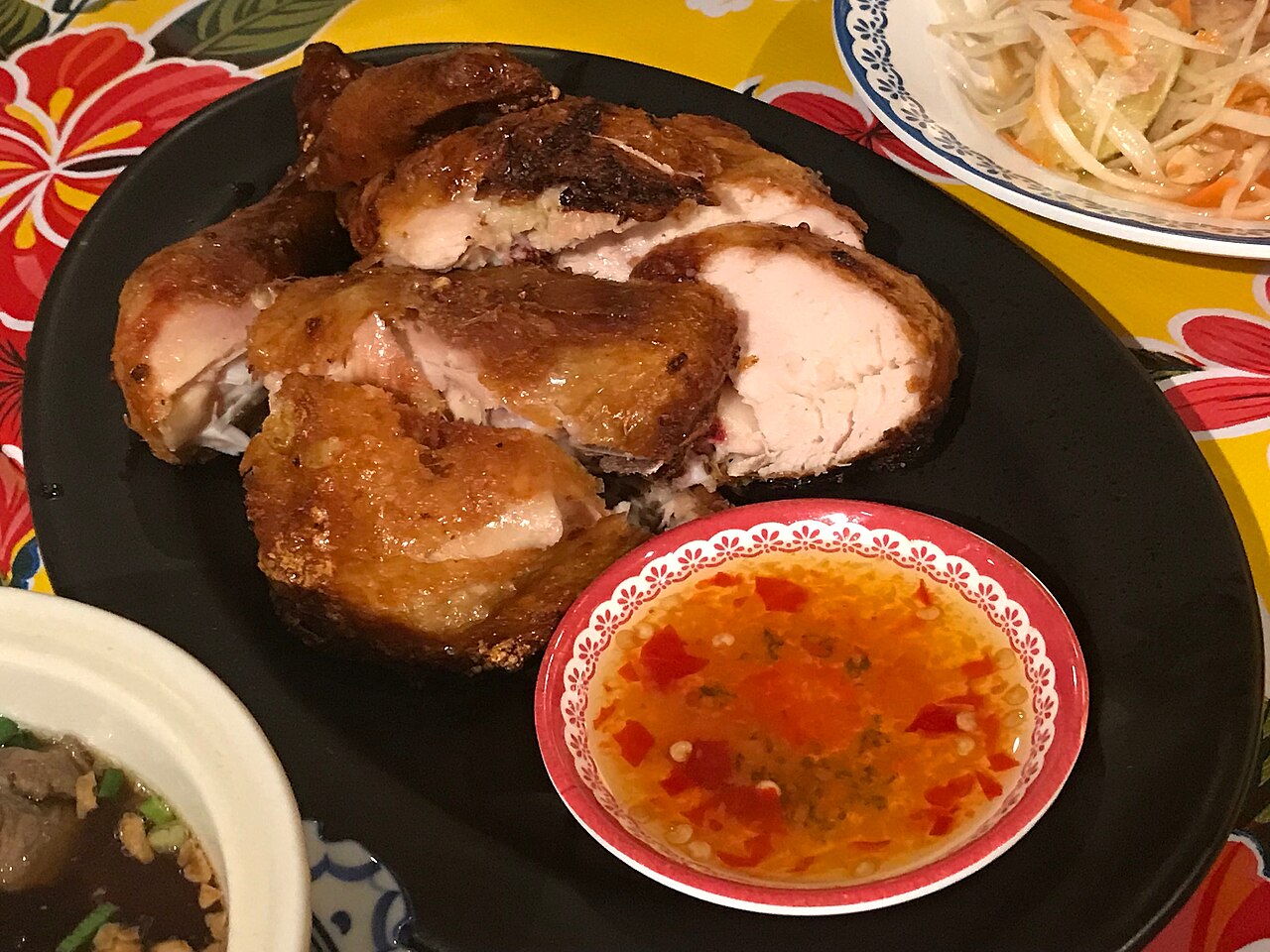 ไก่ย่างหั่นชิ้นบนจานดำพร้อมน้ำจิ้มแจ่วและส้มตำ อาหารอีสานริมทางแบบไทย