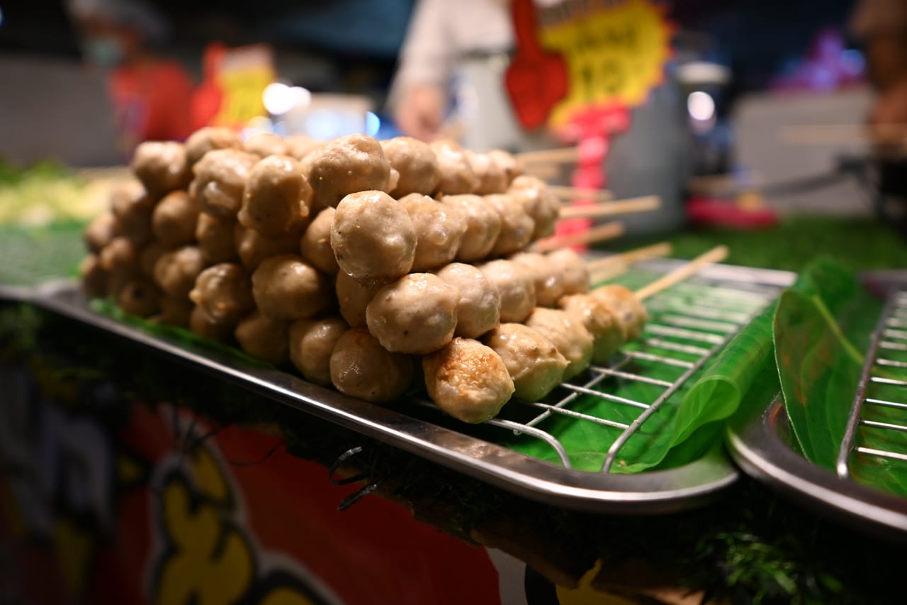ลูกชิ้นปิ้งเสียบไม้บนตะแกรงร้านในตลาดโต้รุ่ง ปูด้วยใบตอง มีป้ายราคาด้านหลัง อาหารปิ้งย่างริมทางแบบไทย