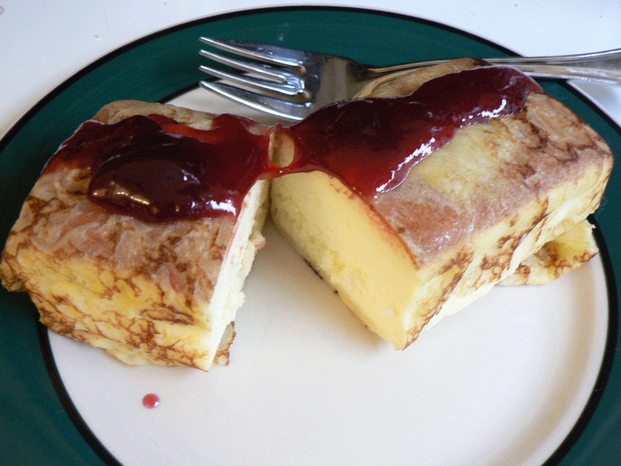 Western-style Kobe dessert — sponge cake slice cut open revealing a pale custard filling, topped with deep-red berry sauce, served on a white plate with a green rim