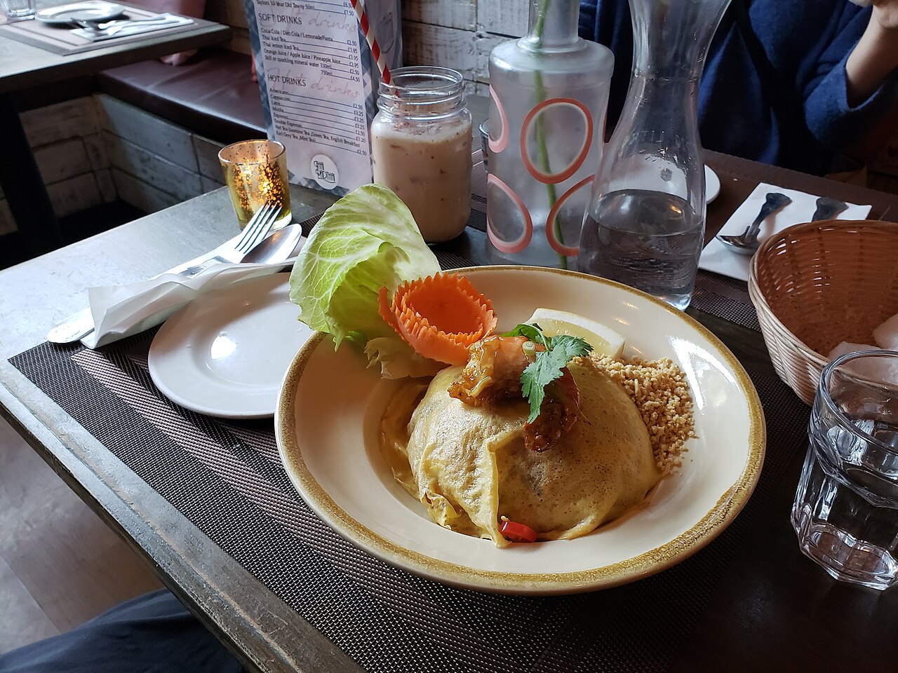 A plate of pad thai wrapped in a thin egg, topped with crushed peanuts, a lime wedge, fresh vegetables and a prawn, a popular made-to-order dish