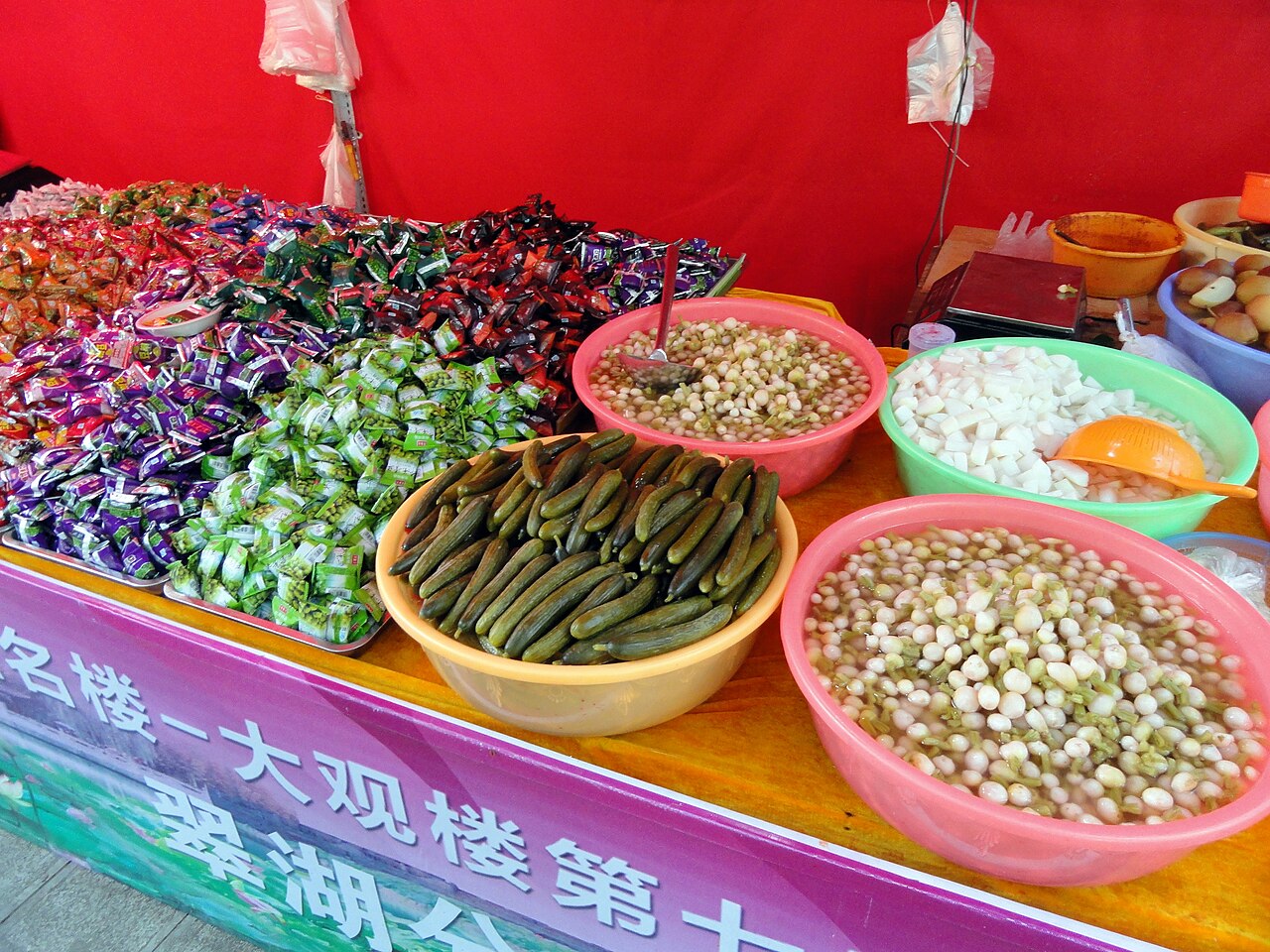 แผงขนมและของกินในตลาดคุนหมิง — กะละมังใส่ถั่วต้ม มะตูมเชื่อม ของหมักผัก และของหวานพื้นเมืองยูนนานวางเรียงบนแผงไม้ ป้ายภาษาจีน