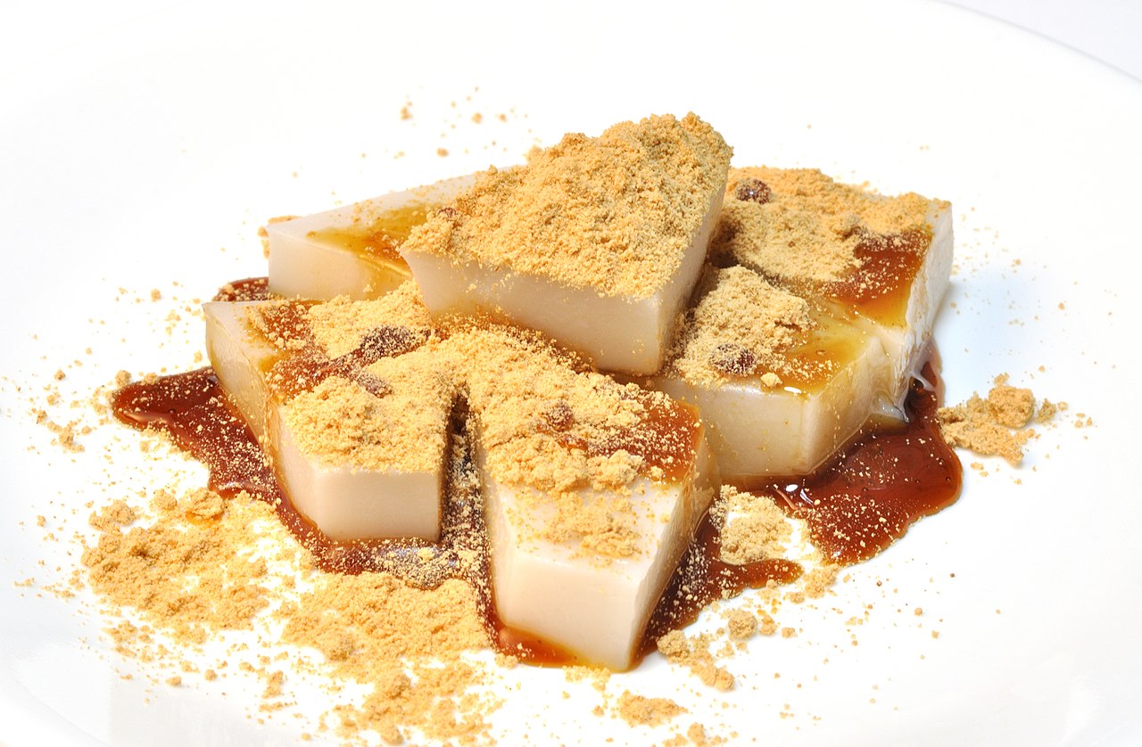 Yoshino kuzumochi: translucent white kudzu-starch jelly cut into thick squares, drizzled with dark kuromitsu brown-sugar syrup and generously dusted with golden kinako soybean flour, on a white plate