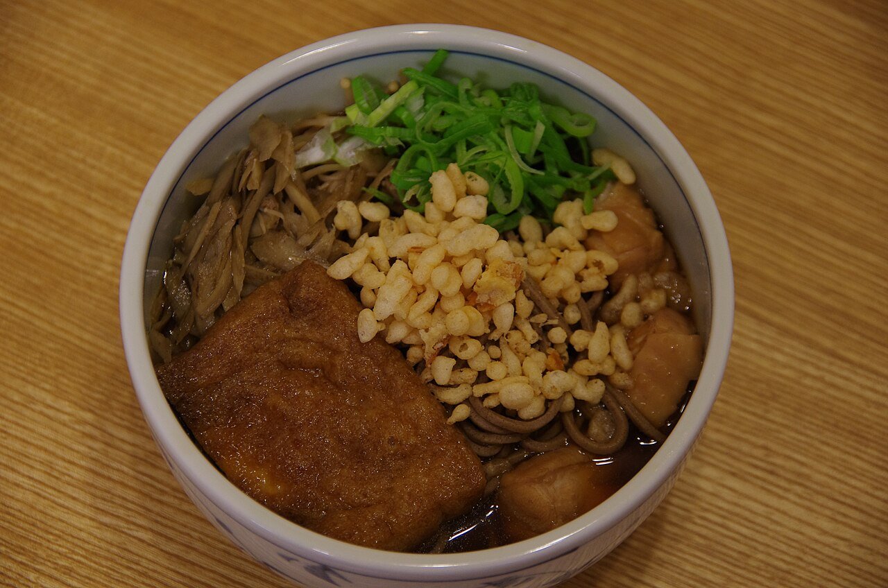 Nikko soba, brown buckwheat noodles in hot broth topped with wild mountain vegetables, fried tofu, tempura crumbs and green onion in a blue-and-white bowl
