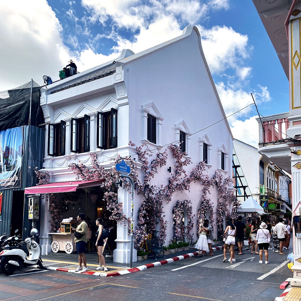 ตึกแถวชิโน-โปรตุกีสมุมถนนในภูเก็ตเมืองเก่า อาคารสีขาวประดับซุ้มดอกไม้ มีคาเฟ่และรถเข็นขายของริมทาง ผู้คนเดินเที่ยวถ่ายรูป