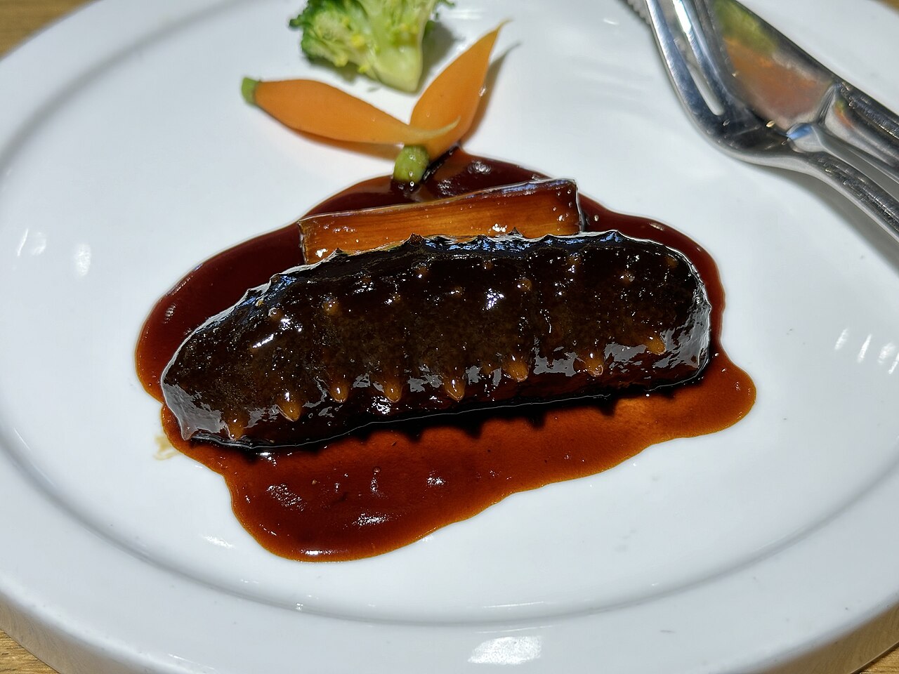 葱烧海参 braised sea cucumber with scallion, a glossy dark sea cucumber resting on a scallion segment in a rich brown soy-sugar sauce on a white plate