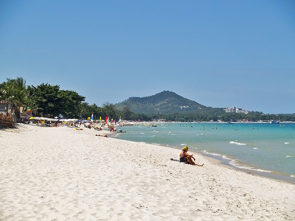 หาดเฉวง เกาะสมุย ทรายขาวโค้งยาว น้ำทะเลใส คนเล่นน้ำและร่มชายหาดเรียงตามแนวต้นไม้
