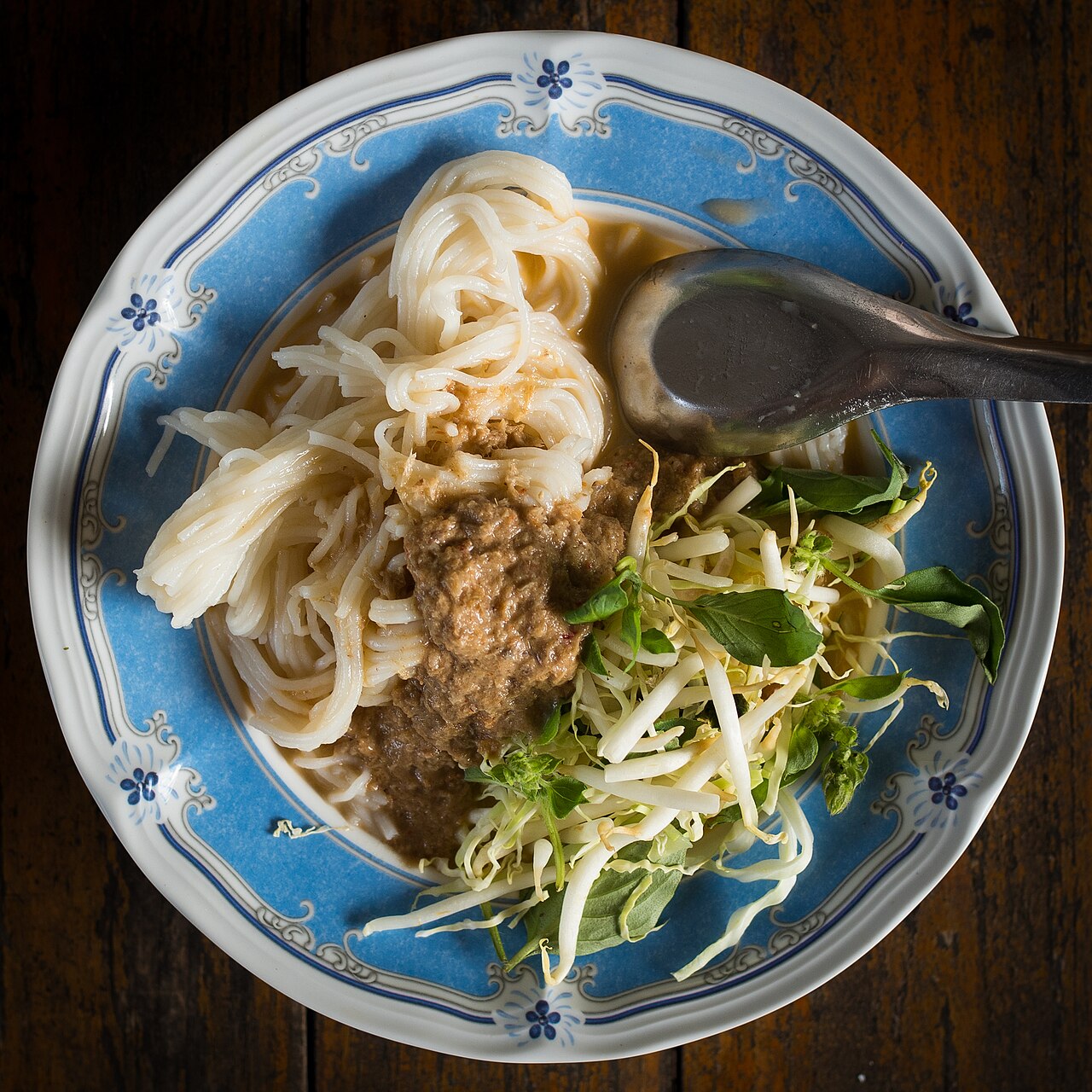 A blue-and-white plate of khanom jeen rice noodles under a thick fish curry, with bean sprouts and fresh herbs