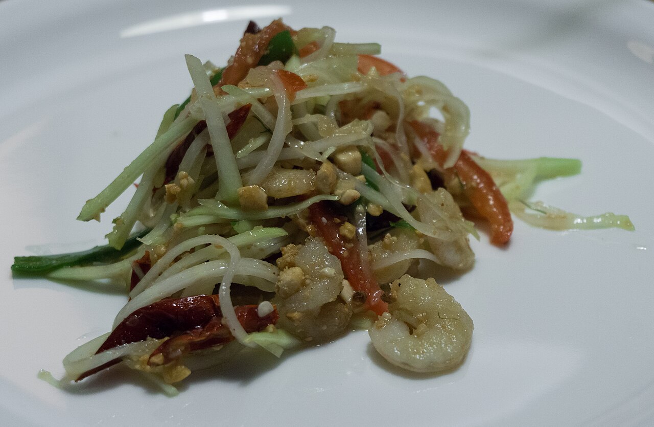A white plate of som tam papaya salad with fresh prawns, tomatoes, dried chillies and peanuts
