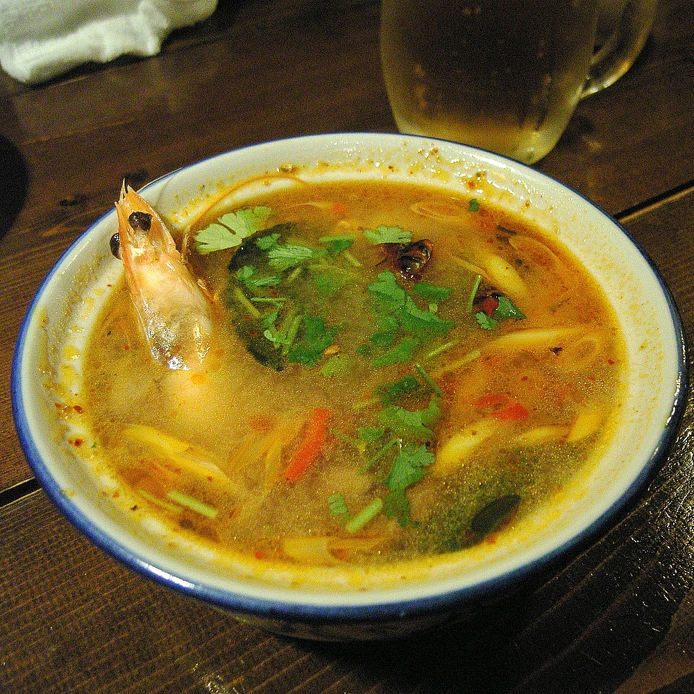 A hot bowl of clear tom yum goong topped with coriander, a prawn head rising from the broth