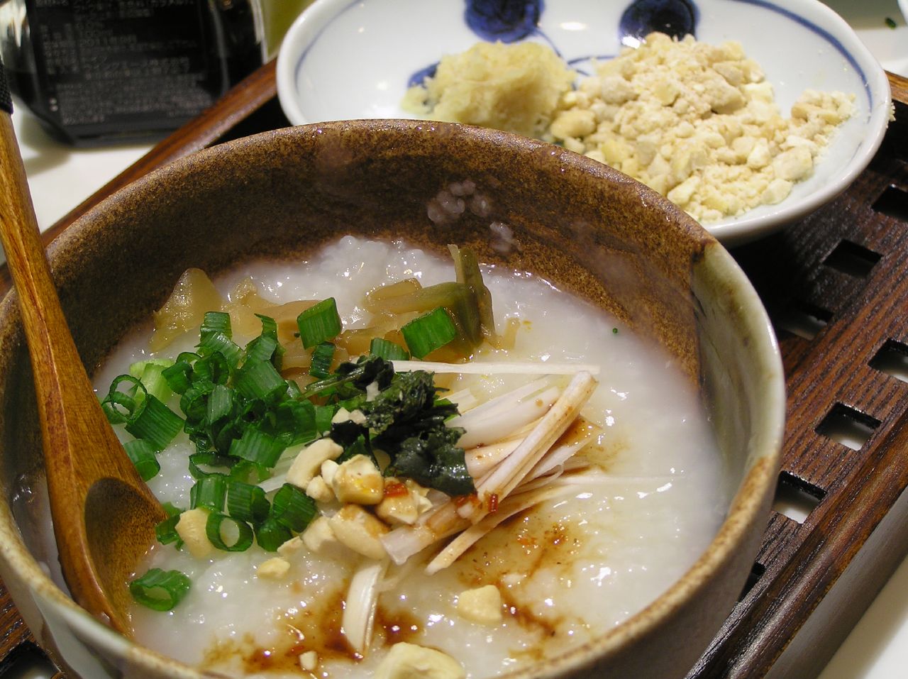 A bowl of Chinese rice congee in a clay bowl topped with scallions and meat, served with side dishes on a wooden tray, a Hainan-style breakfast