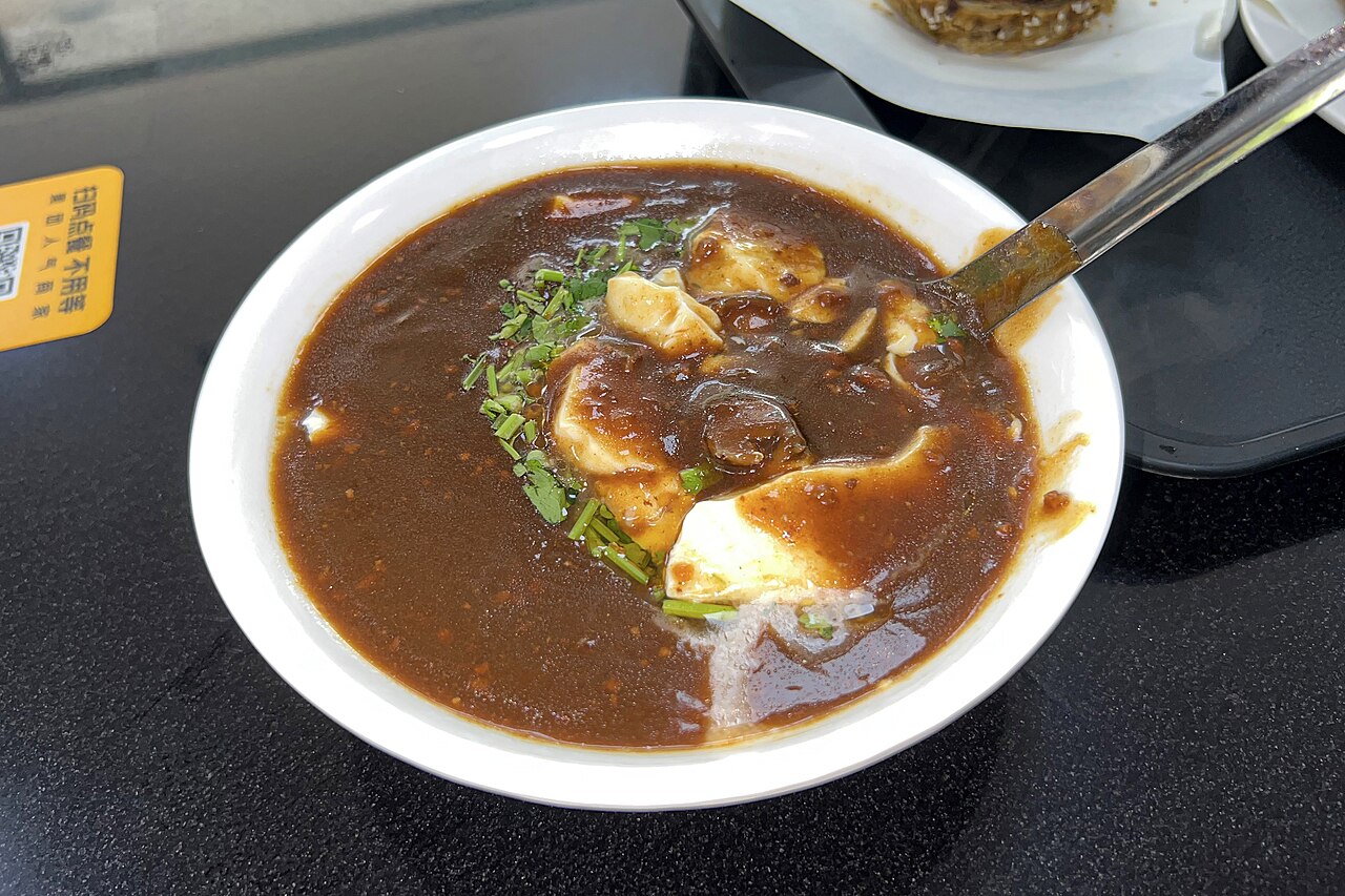 Doufunao — soft white tofu pudding in a dark brown savoury halal gravy scattered with coriander, in a white bowl