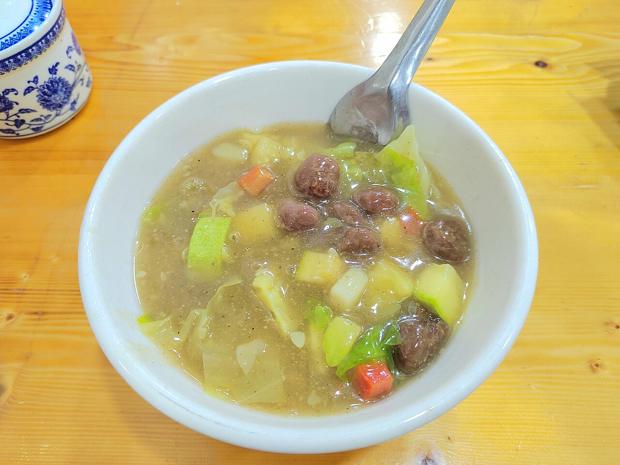 A bowl of Xi'an hulatang — thick brown peppery soup with mutton meatballs, diced potato, cabbage and carrot, in a white bowl on a wooden table